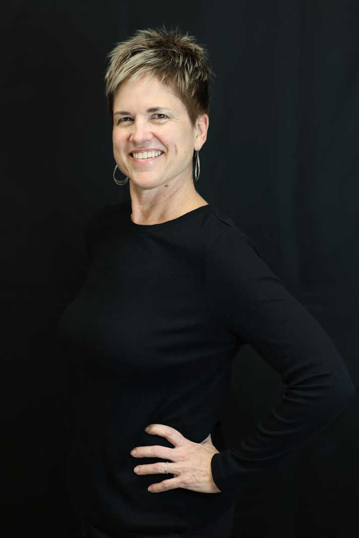 A woman with short pink hair smiling, wearing a black sleeveless top, standing against a plain white wall.