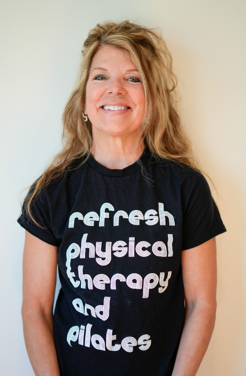 A woman with wavy red hair and blue eyes taking a selfie indoors, smiling, wearing a black top, with wooden and glass window panes in the background.