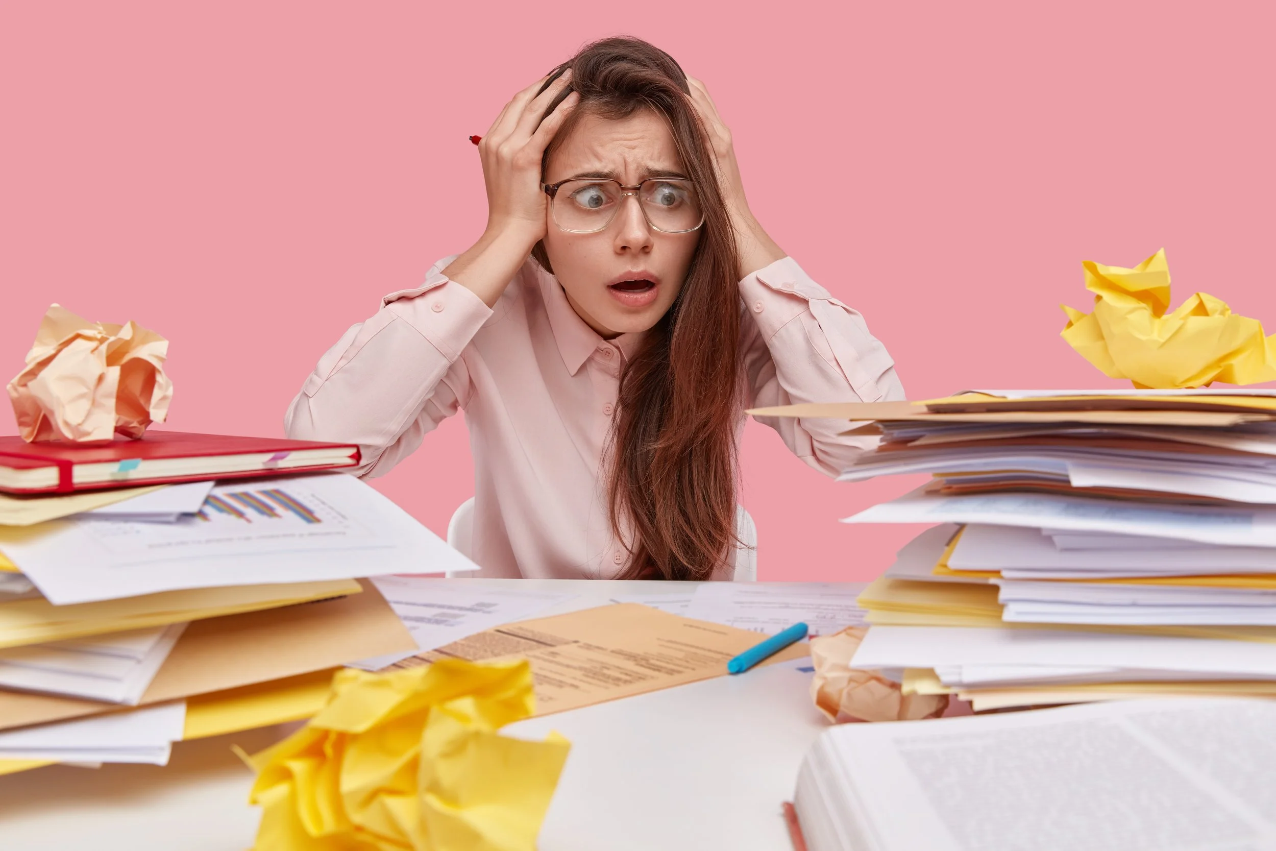 A woman looks overwhelmed and stressed, sitting at a cluttered desk with piles of papers, documents, and crumpled papers, against a pink background.