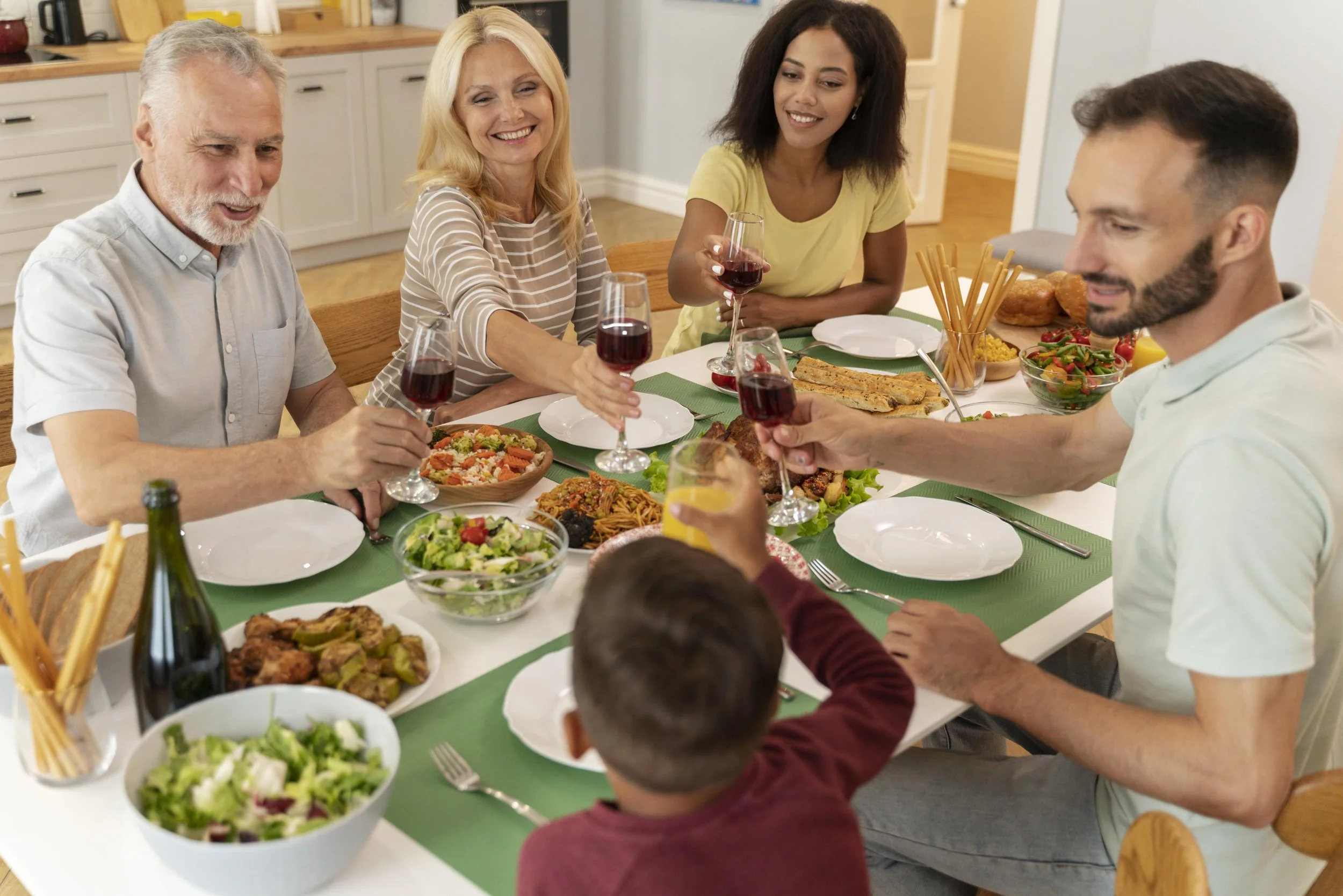 A multi-generational family enjoying a dinner together at a dining table with various foods and drinks, raising glasses for a toast in a home kitchen or dining area.