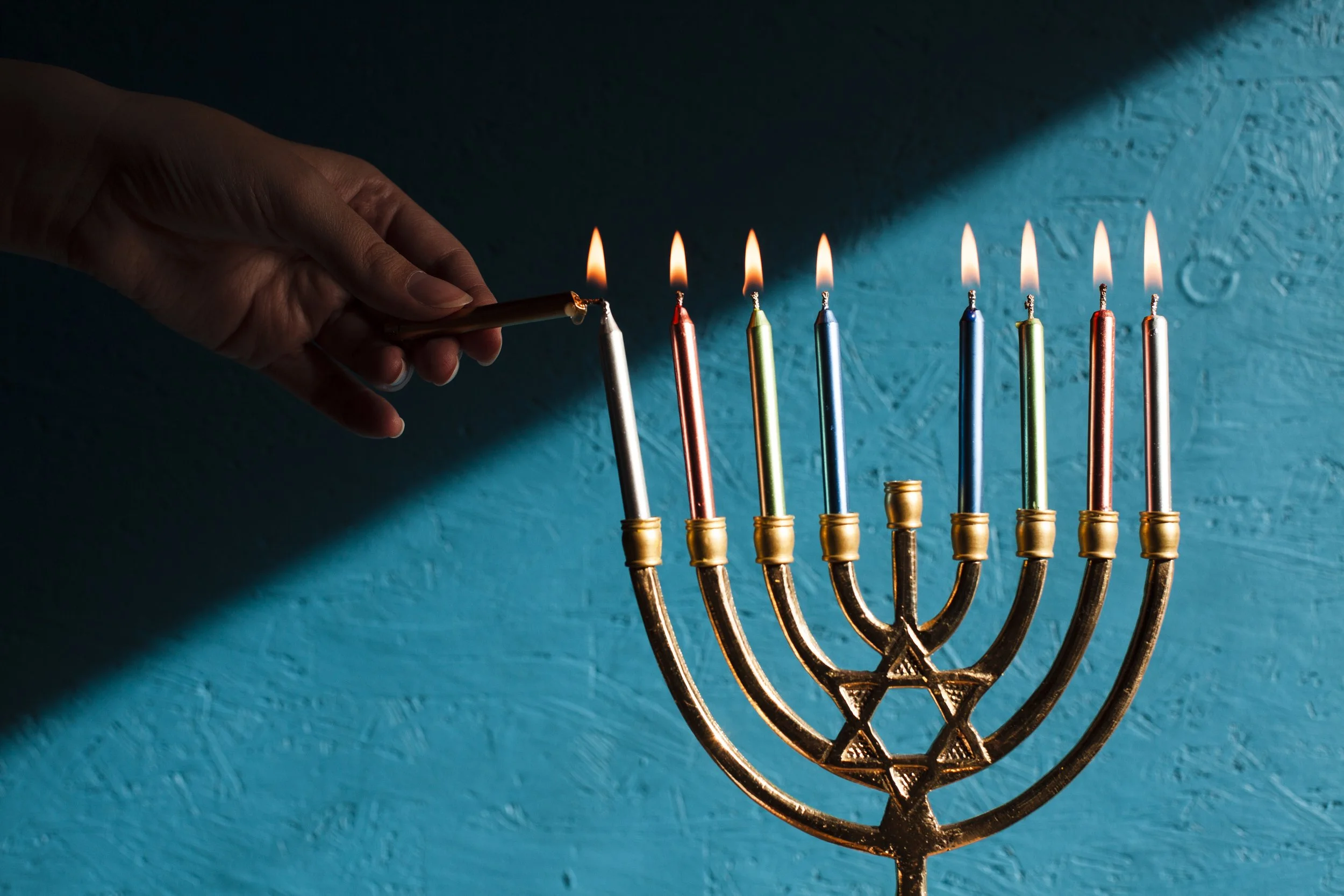 A hand lighting a birthday candle on a menorah with lit candles, against a textured blue wall.