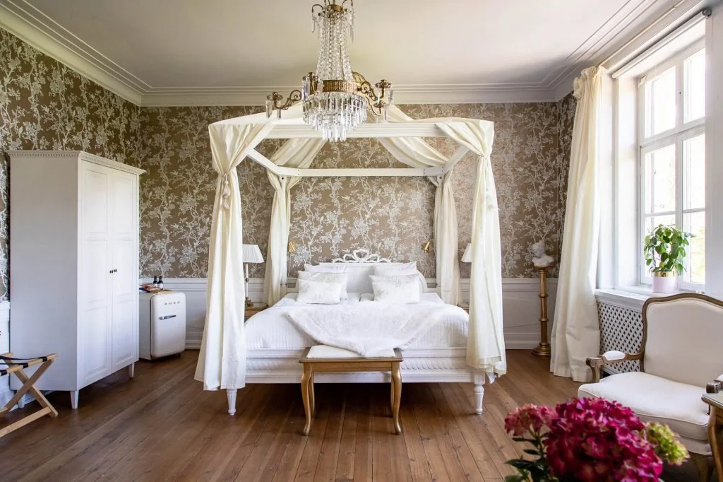 Elegant bedroom with a four-poster bed, chandelier, floral wallpaper, and large window with white curtains.
