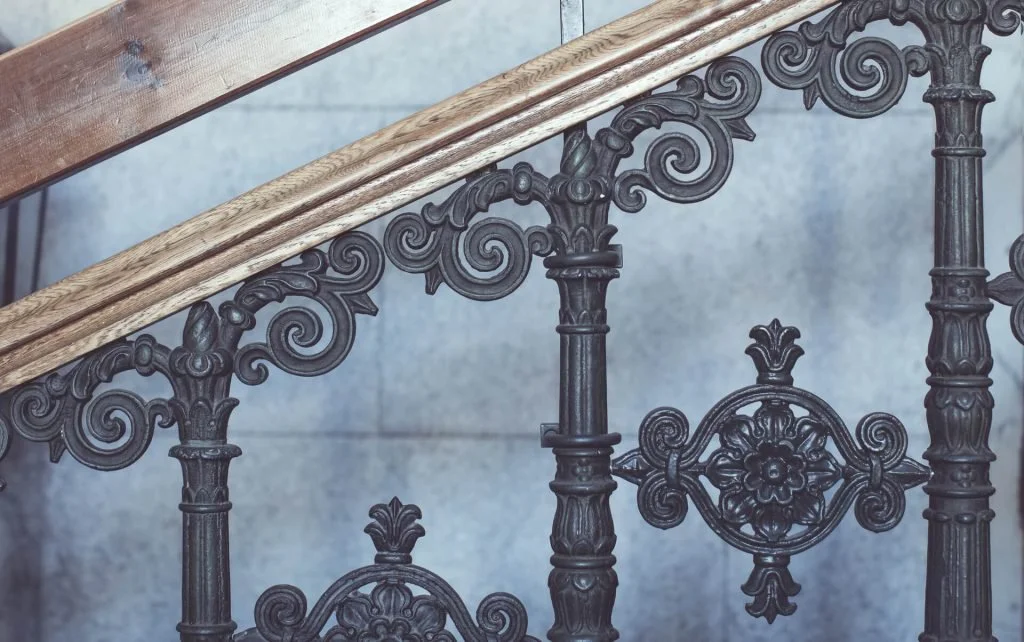 Close-up of a decorative iron railing with intricate floral and scrollwork designs, attached to a wooden handrail.