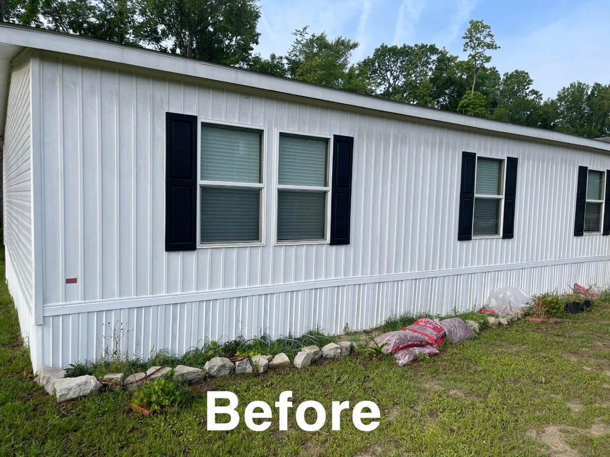 White mobile home with black window shutters, small flower bed with stones and plants, and green grass in front.