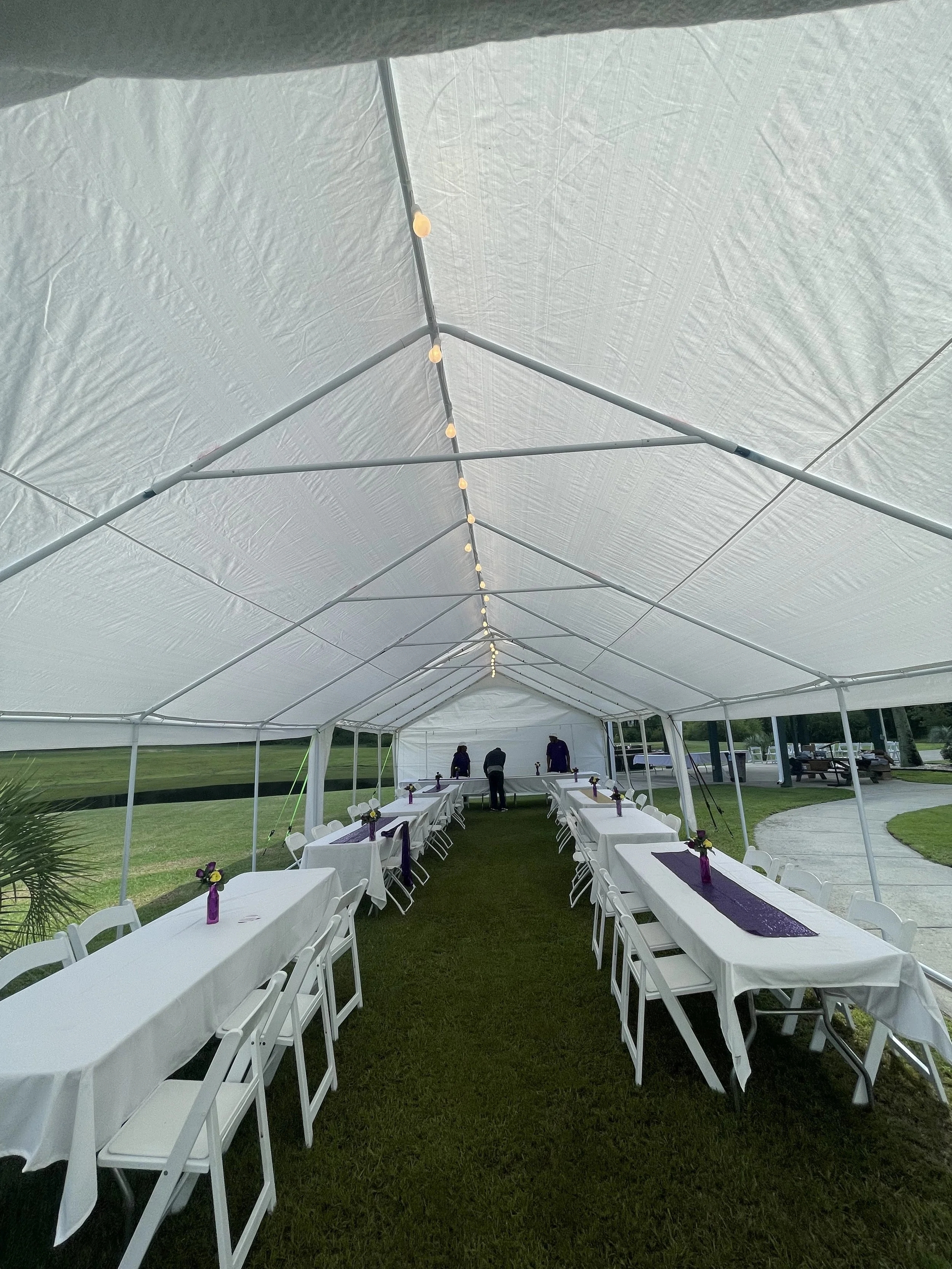 Setup for an outdoor event with white tables and chairs underneath a large white tent, decorated with purple table runners and small flower arrangements, with a grassy area and park-like setting in the background.