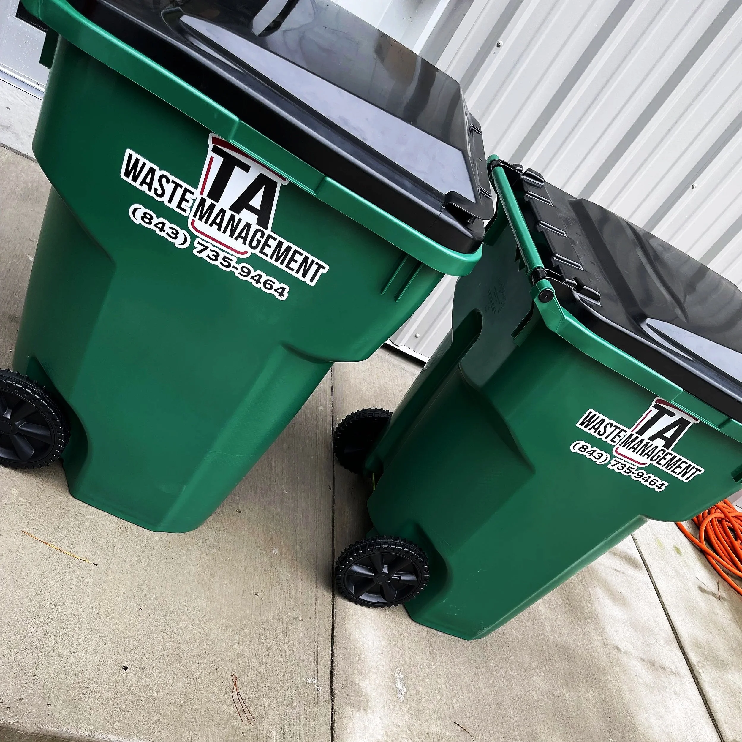 Two green waste management trash bins with black lids and wheels, placed on a concrete surface near a metallic wall.