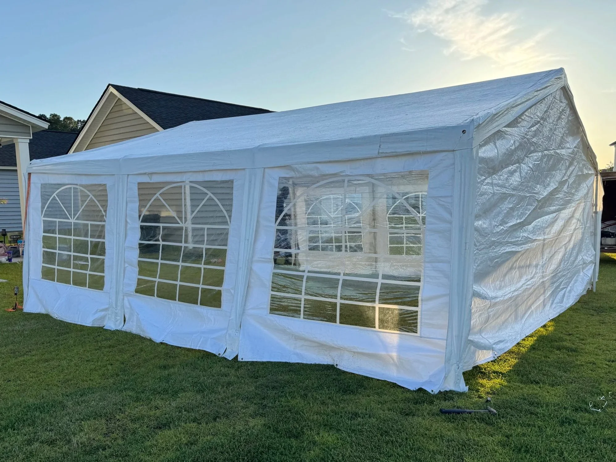 A white outdoor canopy tent set up on a grassy yard in front of neighboring houses, with arch-shaped window panels and partially rolled-up sides, during daytime.