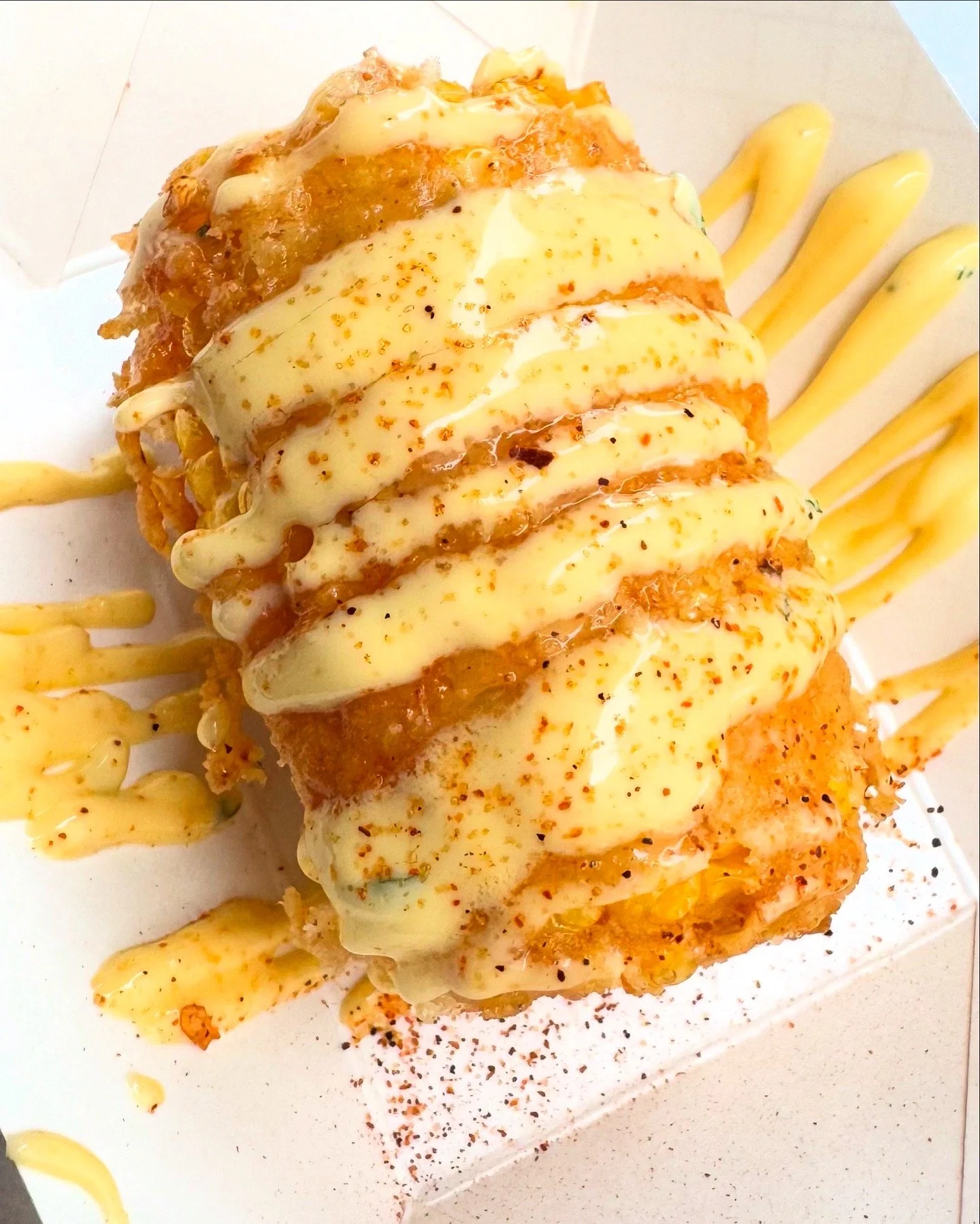 Fried corn topped with creamy sauce, garnished with cajun seasoning, and drizzled with additional sauce, served on a white plate with decorative sauce streaks.