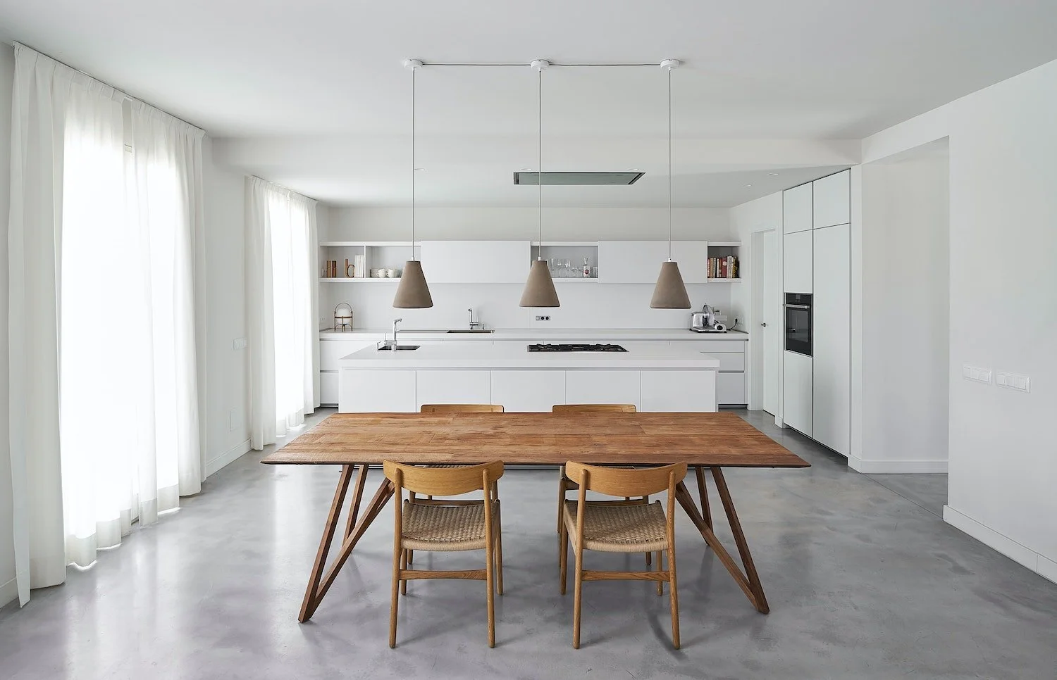 Minimalist white kitchen with a wooden dining table and chairs, pendant lights, and large windows with white curtains.