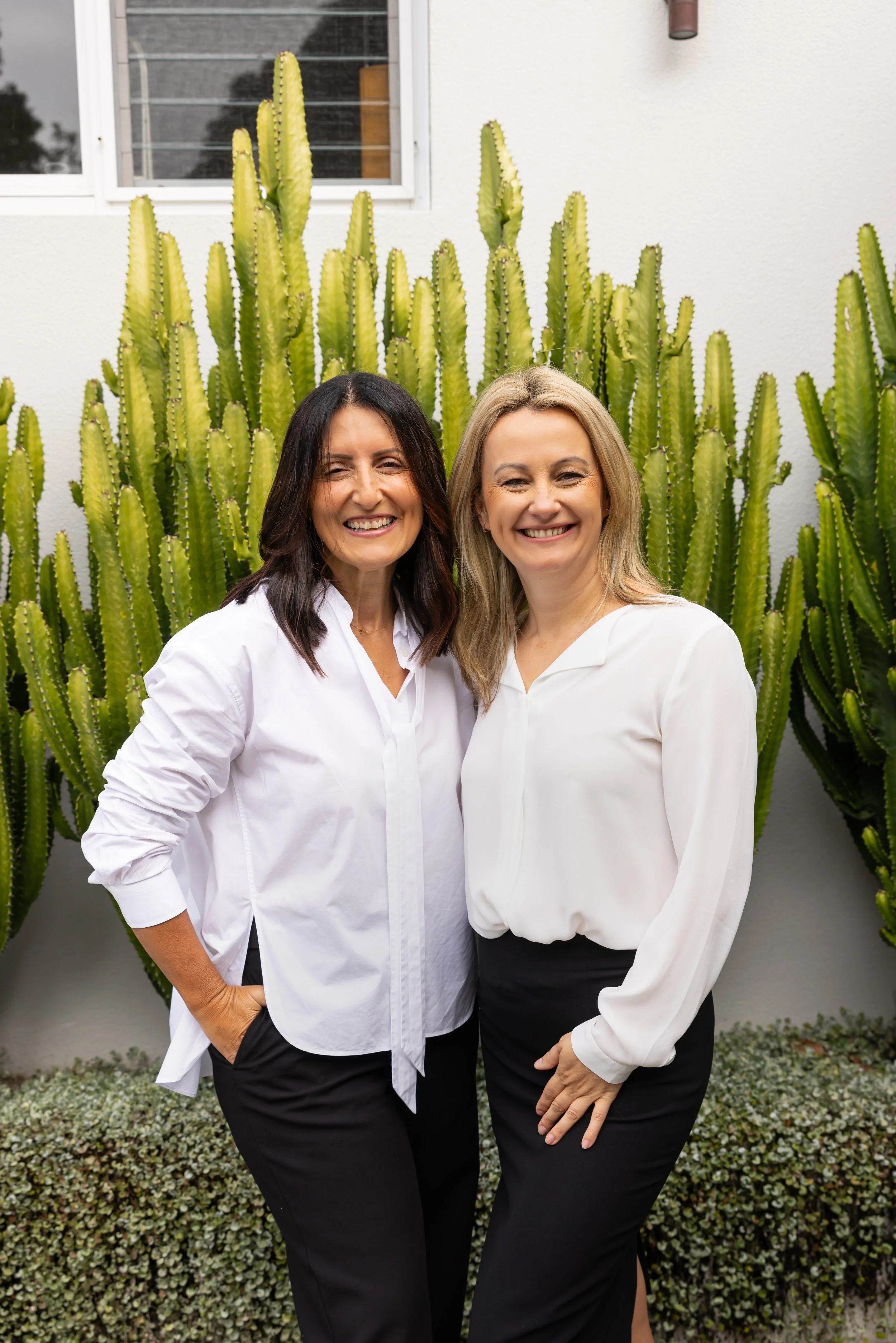 Irene and Nada, founders of IN Property Agents smiling and standing in front of large cactus plants outside a modern home in Sydney Eastern Suburbs.