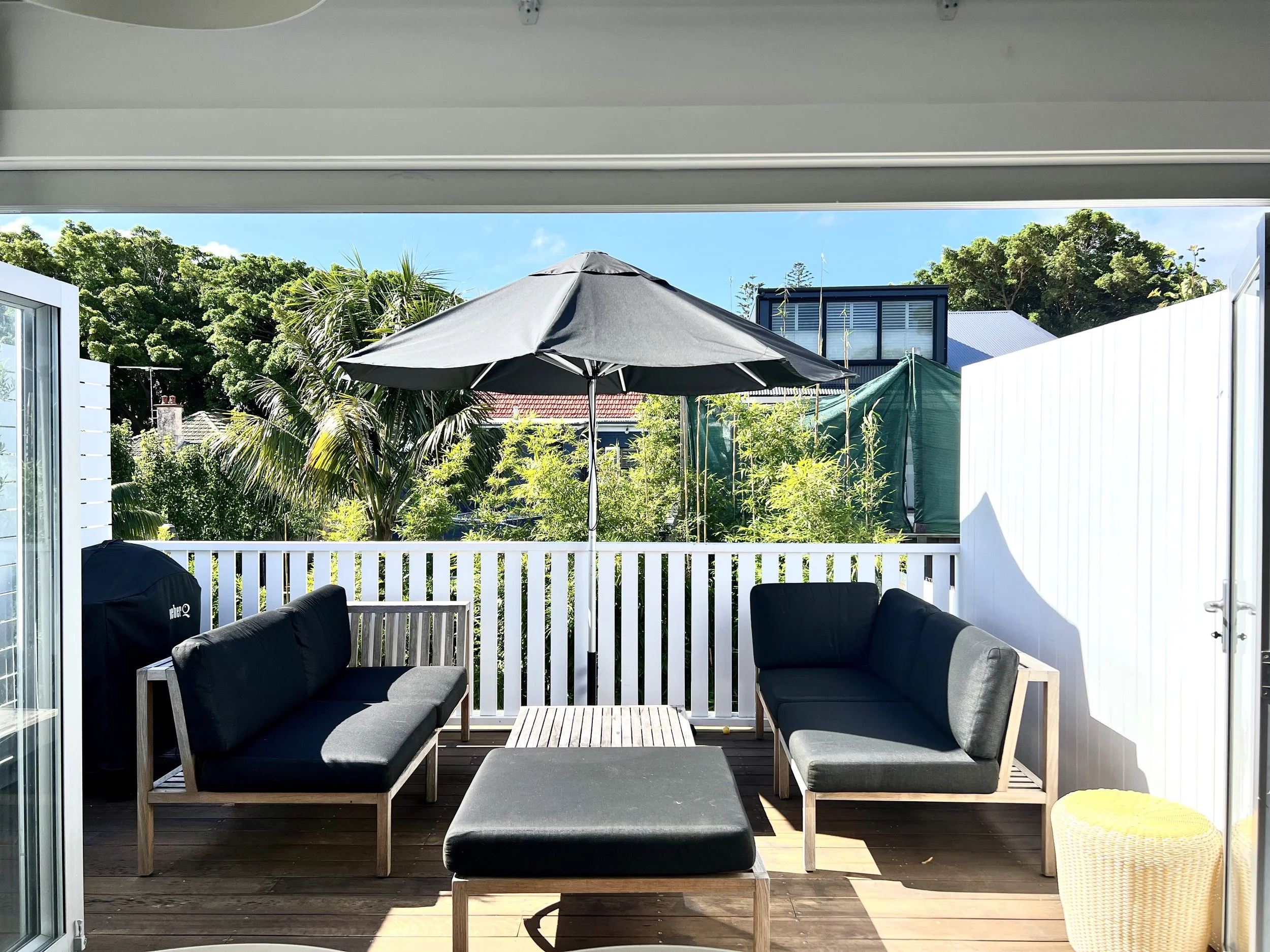 An after photo showing a cozy outdoor patio with black cushioned seating, a small wooden coffee table, an umbrella, and a view of lush green trees and neighboring houses under a bright blue sky.