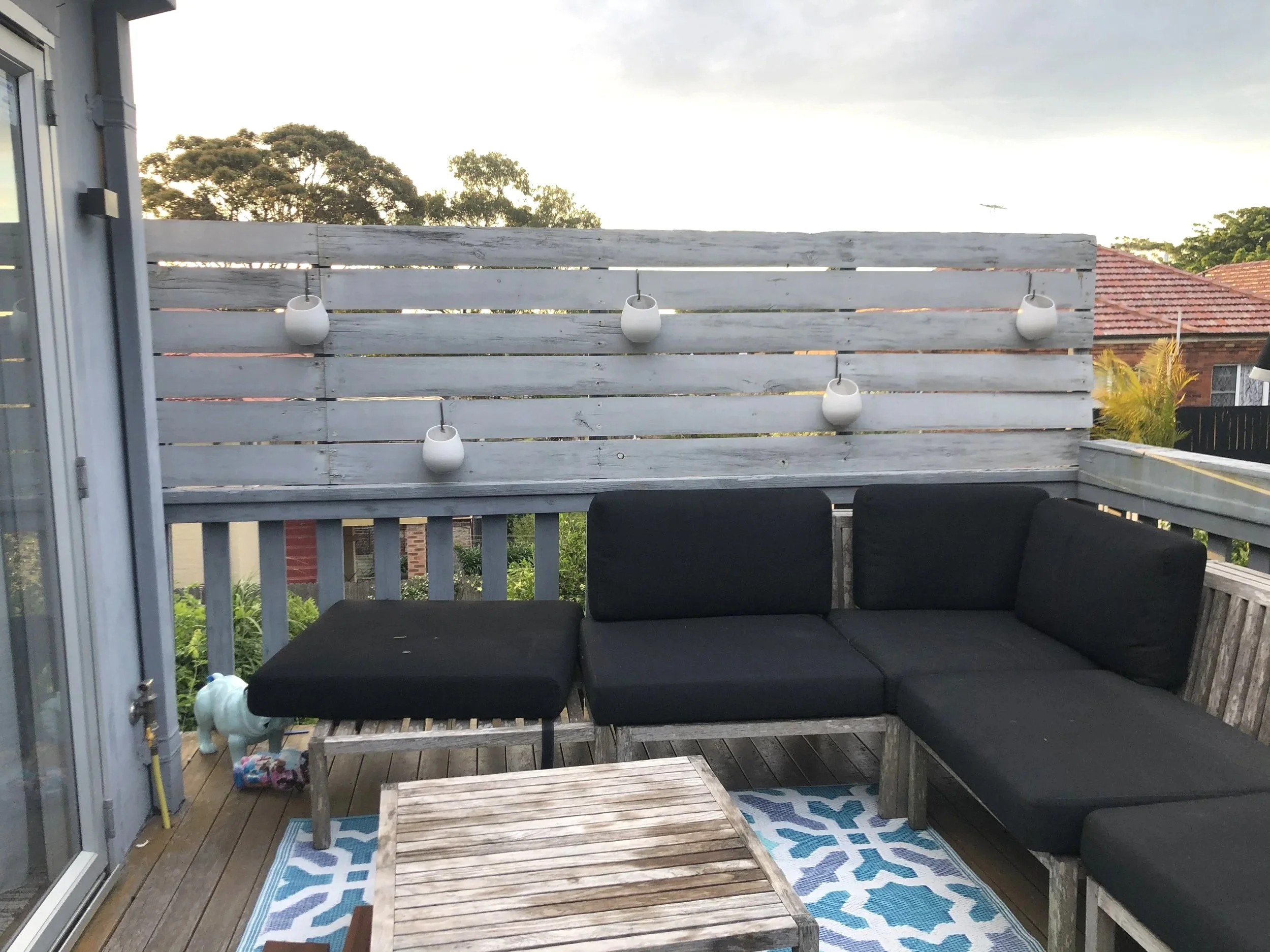 A before photo showing a outdoor balcony with a black cushioned seating area, on a wooden deck, with trees and houses in the background.