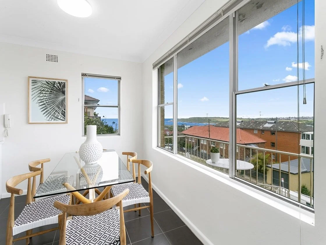 Dining area with a glass table, four wooden chairs with patterned cushions, a white vase, and large windows showing a residential neighborhood and a view of the ocean in the distance.