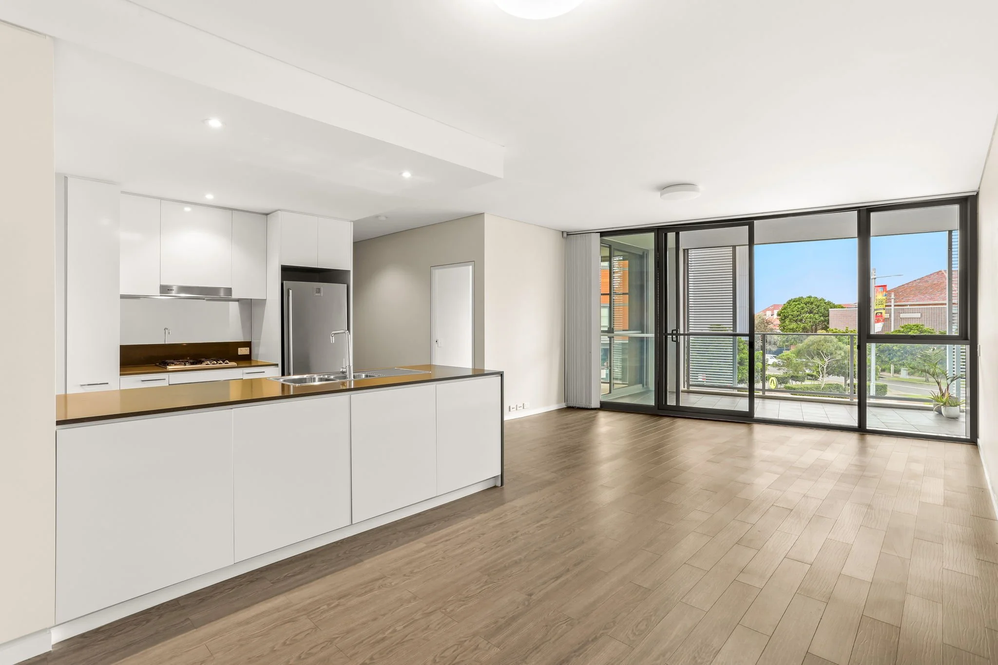 Modern open-concept living space with white kitchen cabinets, a wooden countertop, and large sliding glass doors leading to a balcony with greenery and neighboring buildings.