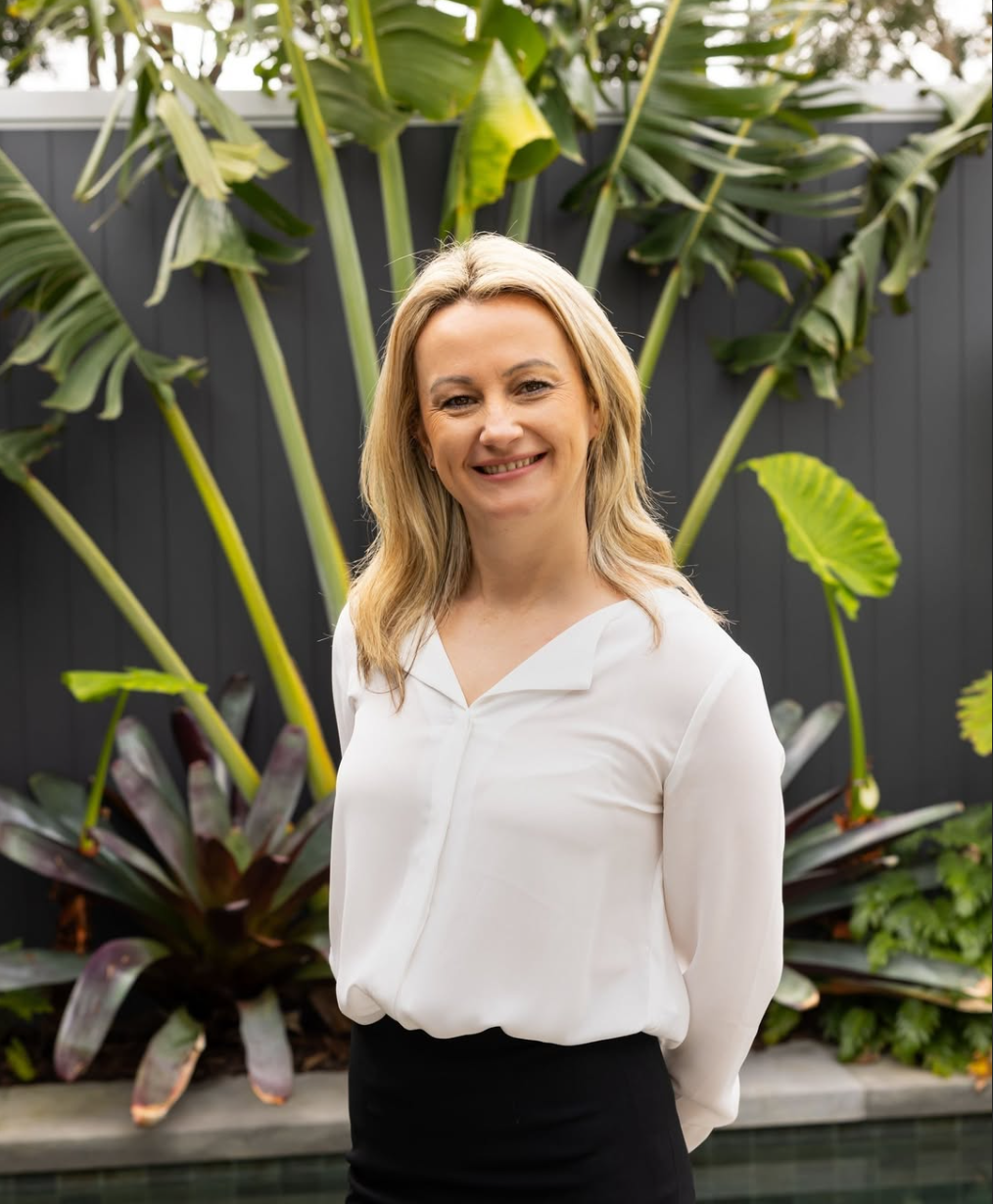 Nada, one of the founders of IN Property Agents standing outdoors in front of lush green tropical plants and a dark fence.
