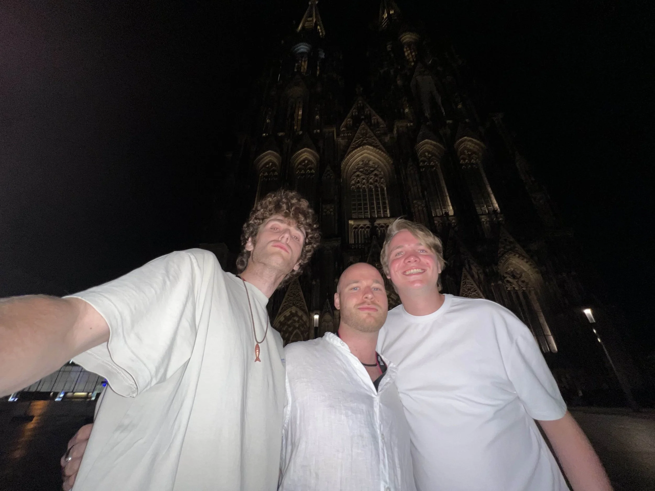 Picture of three handsome men all wearing white clothes and mogging the camera. They are standing in front of the cathedral of Cologne, Germany