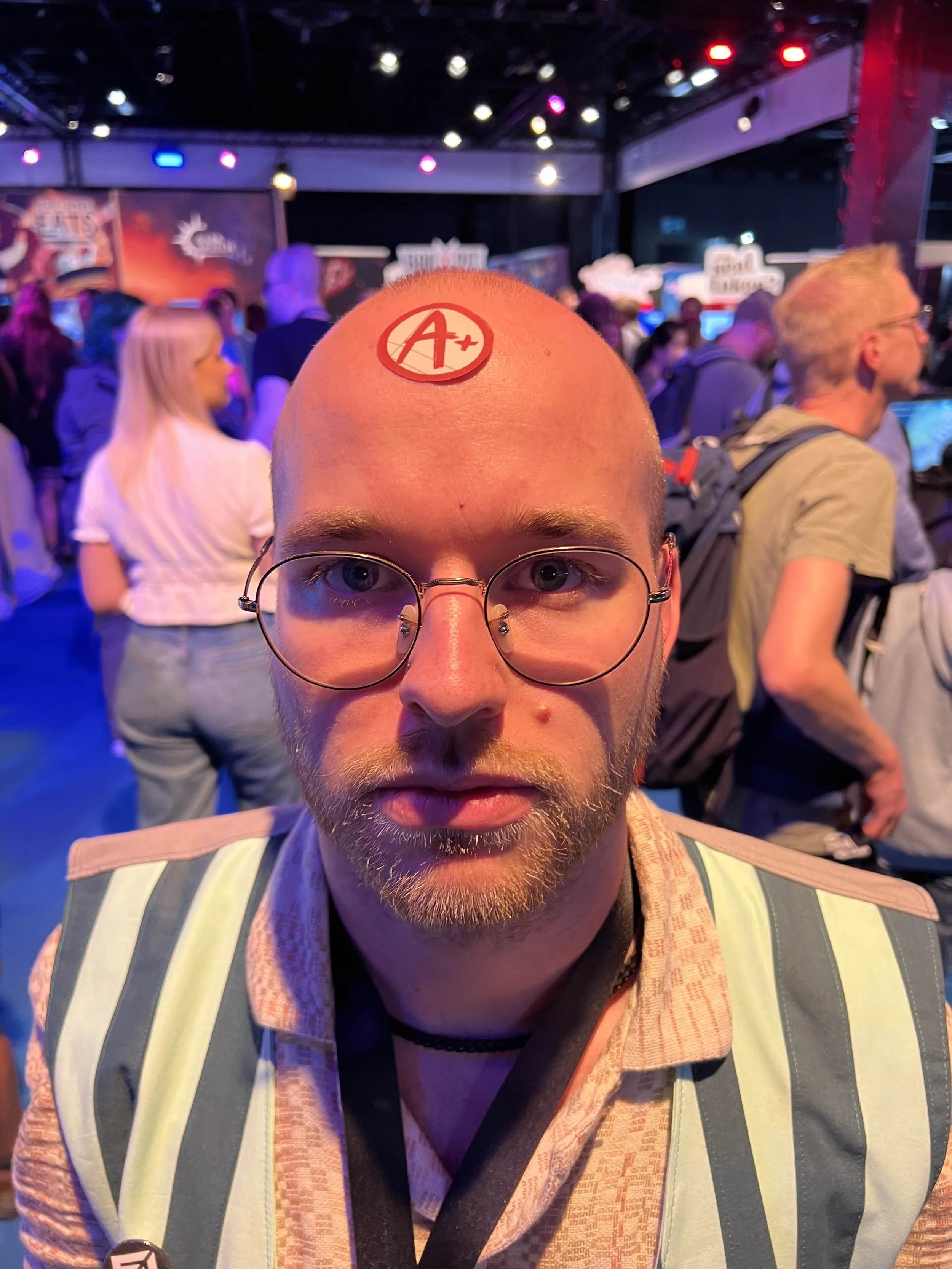 A bearded man standing in the Indie Arena Booth of Gamescom 2025, wearing a vest and with a sticker on his forehead.