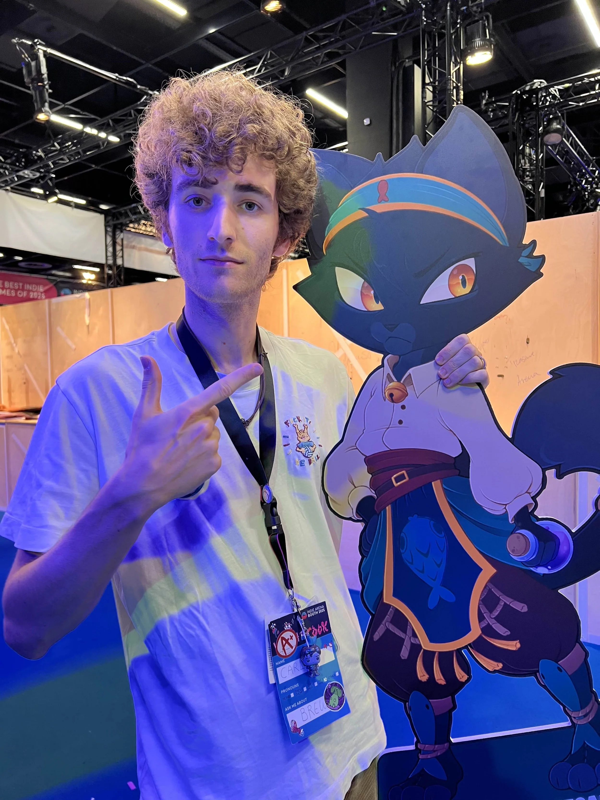 A curly-haired man standing in the Indie Arena Booth of Gamescom 2025 holding a cut-out stnad of Cinna, one of the main characters of the roguelite game Brew, which came out on Steam, November 2025.