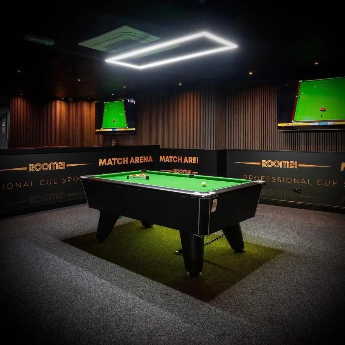 A black pool table with green felt in a dimly lit room, with two white cue balls and a box of cue chalk on the table. Two television screens showing the same pool game are mounted on the dark wood-paneled walls. The room has branding for "ROOM2" and "MATCH ARENA," and the ceiling features modern rectangular light fixtures.