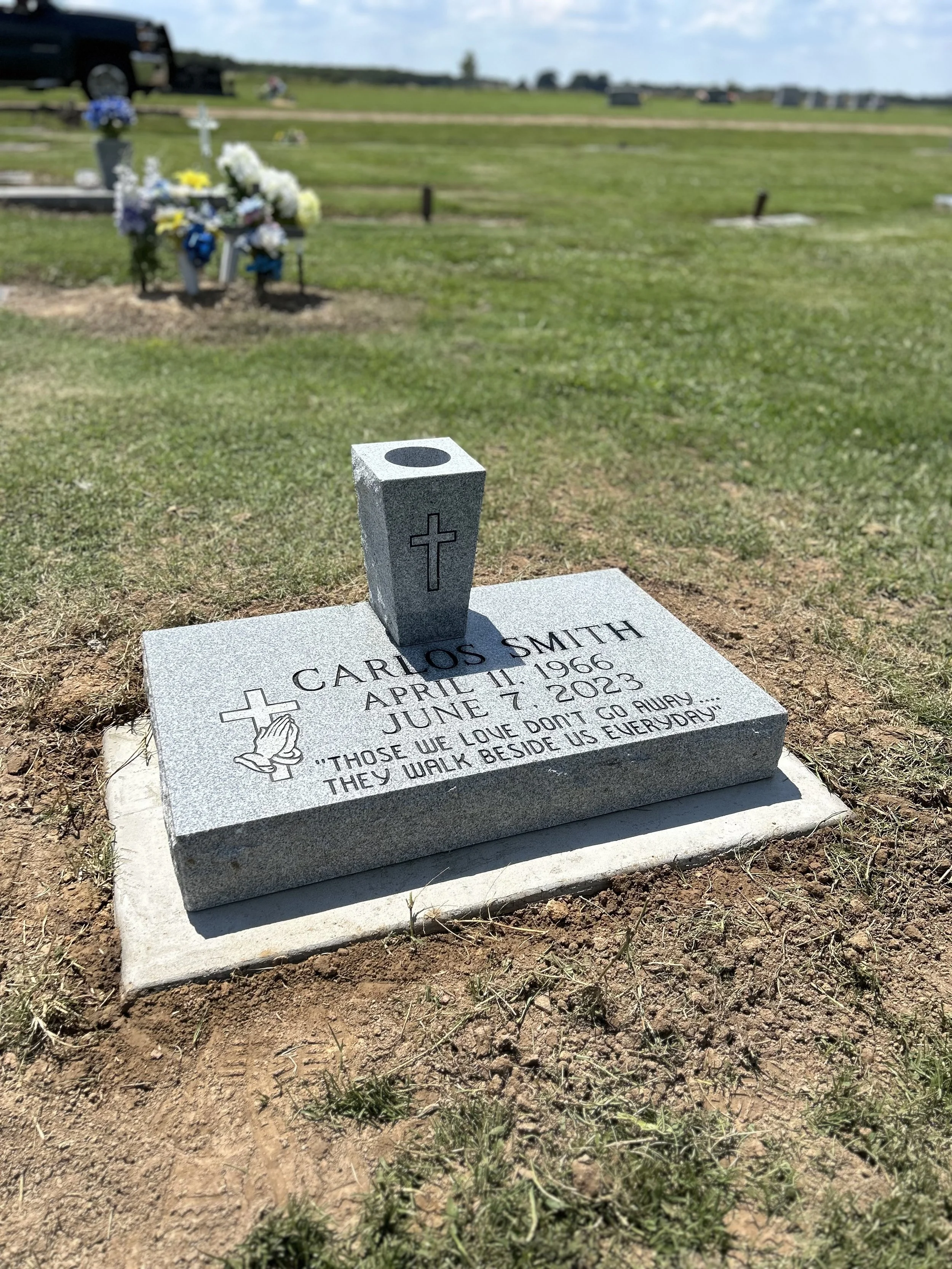A gravestone at a cemetery with the name Carlos Smith, born April 11, 1966, died June 7, 2023. The gravestone has a cross and praying hands engraved, along with the quote: 'Those we love don't go away, they walk beside us every day.' In the backgroun
