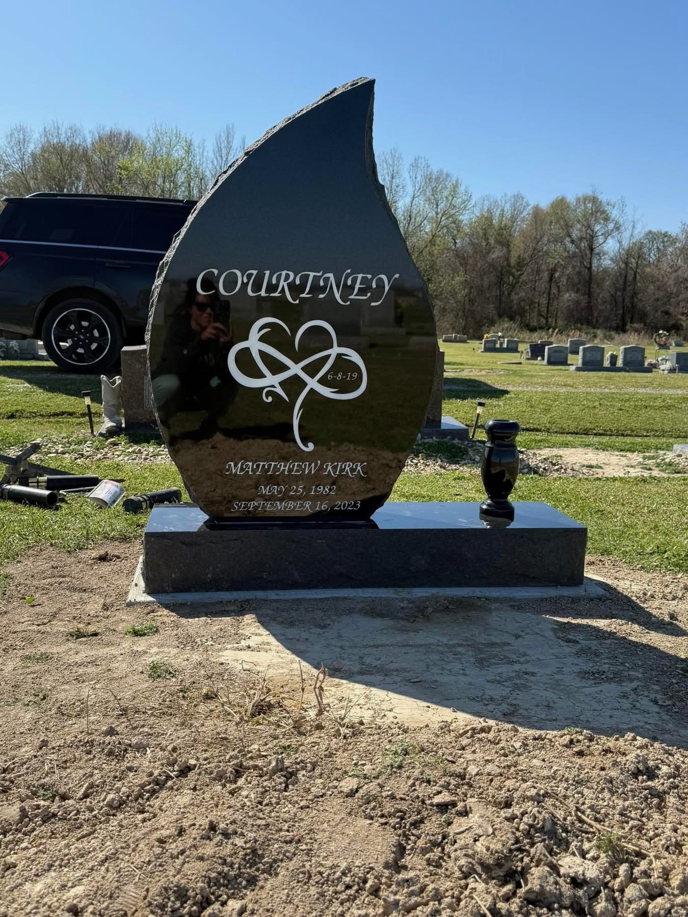 A polished black headstone at a cemetery with the name Courtney engraved, along with a heart-shaped decor and the name Matthew Kirk, birth date May 25, 1982, and death date September 16, 2023. The headstone is shaped like a flame or a leaf, placed on