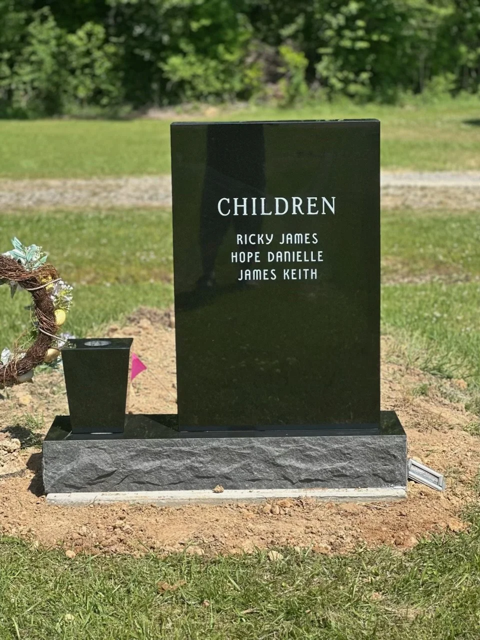 A black granite headstone with the word 'CHILDREN' engraved at the top. Below it are the names Ricky James, Hope Danielle, and James Keith. The headstone is placed on a gray stone base in a grassy area with trees in the background.