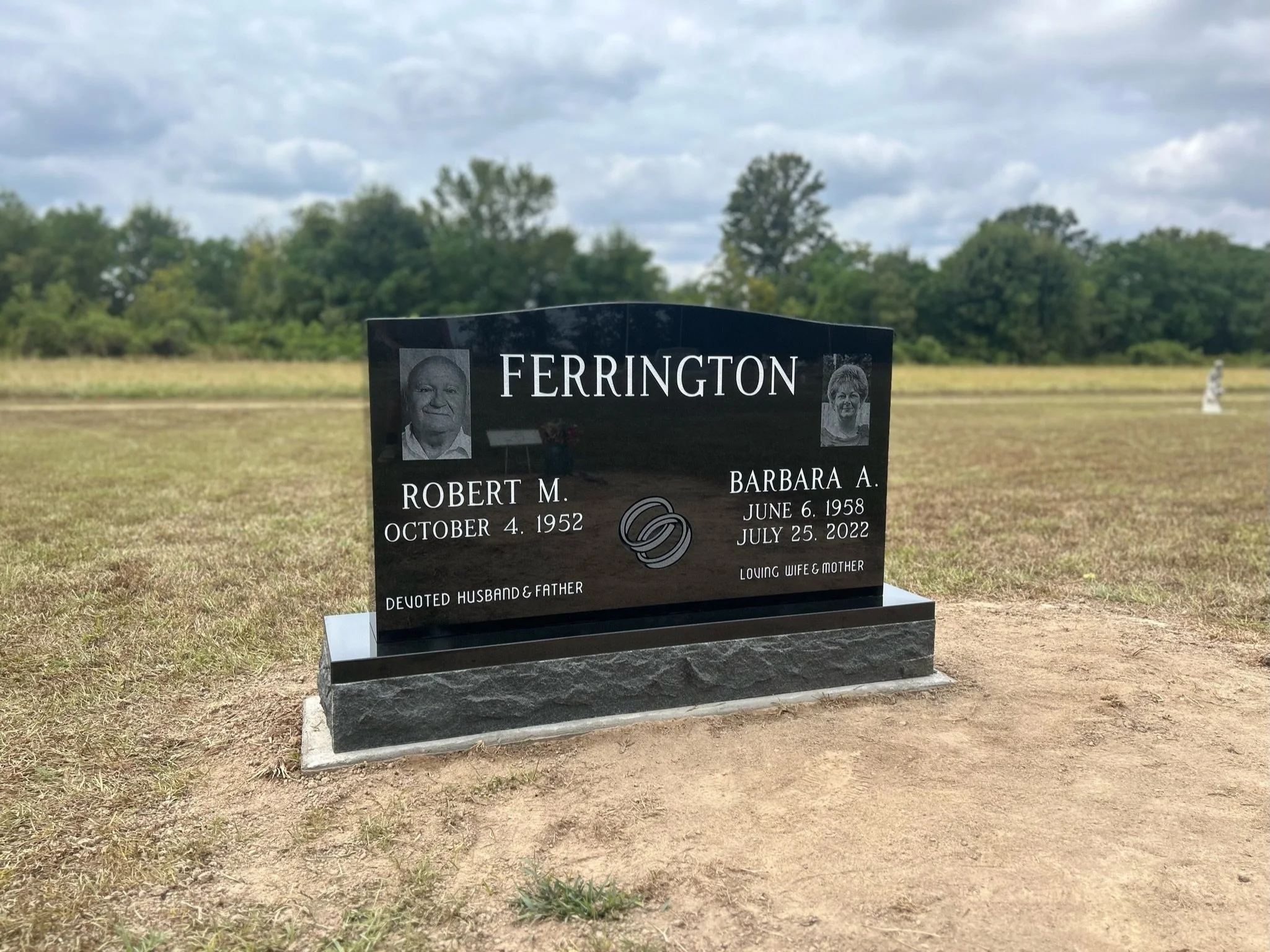 Black granite tombstone with photographs, names, and dates for Robert M. Ferrington (October 4, 1952) and Barbara A. Ferrington (June 6, 1958 - July 25, 2022), set in a grassy cemetery with a distant tree line and cloudy sky.