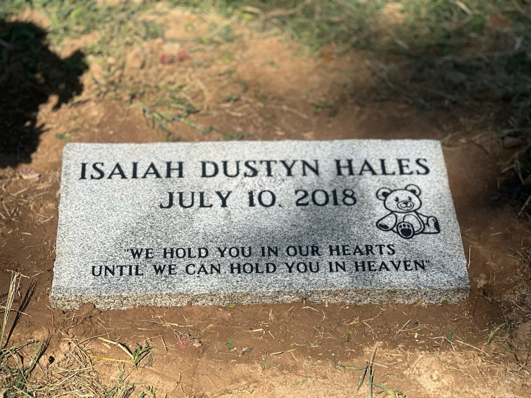 Grave marker for Isaiah Dustyn Hales with the date July 10, 2018, featuring a drawing of a teddy bear and the quote 'We hold you in our hearts until we can hold you in heaven'.