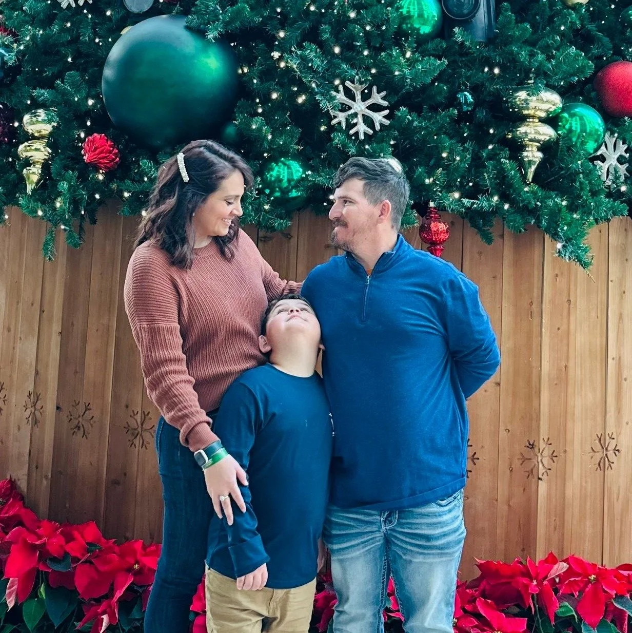 A family of three, a woman, a man, and a boy, standing in front of a decorated Christmas tree with red poinsettias at the base. The woman and man are smiling at each other, while the boy looks upward.