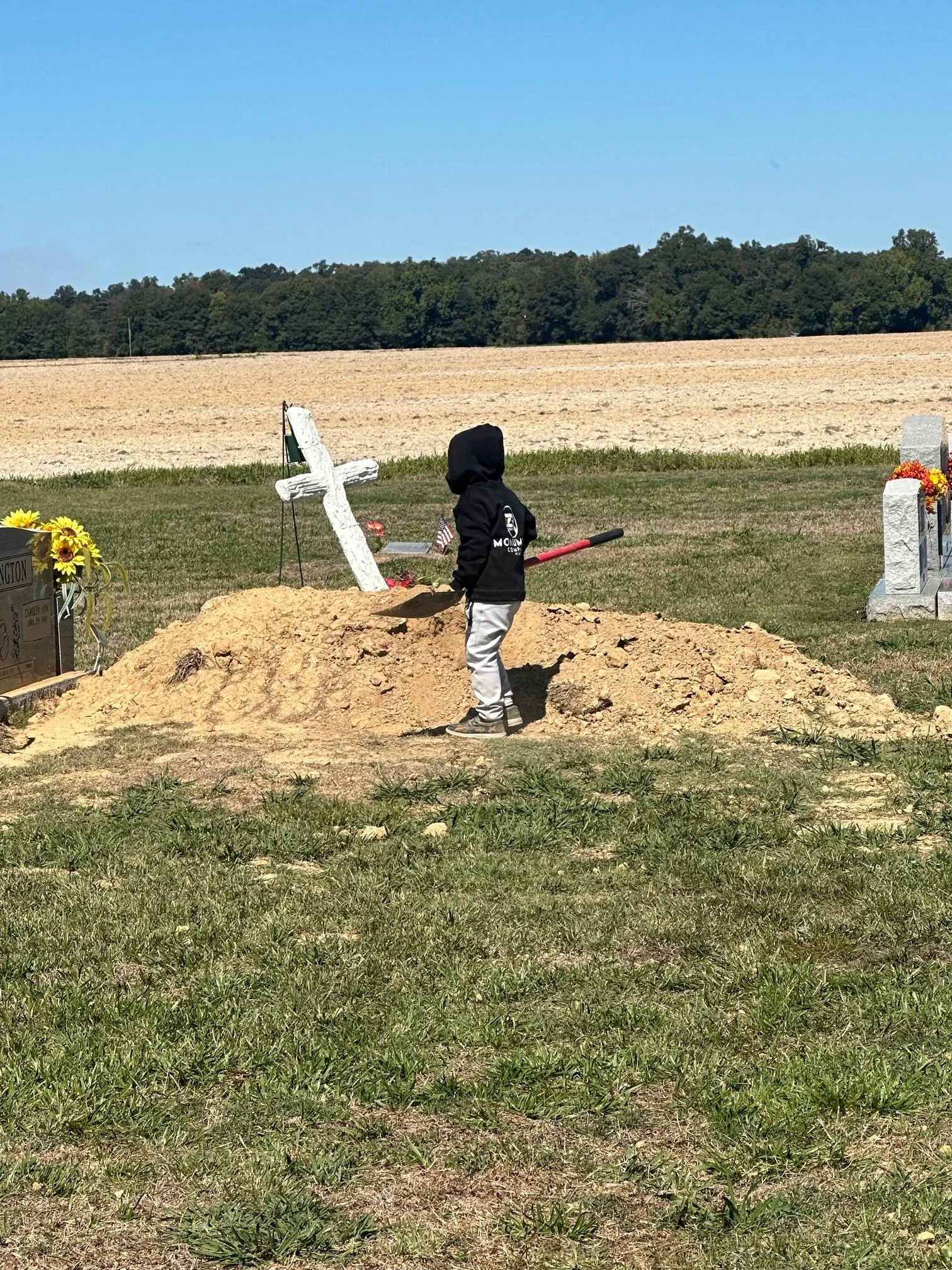 A child in a black hoodie and gray pants shoveling dirt onto a grave at a cemetery, with a white cross and flowers nearby, under a clear blue sky.