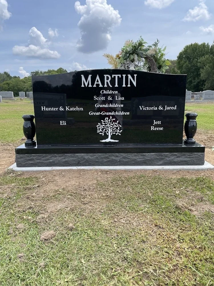 A black granite gravestone with the surname 'Martin' engraved at the top, featuring names of children, grandchildren, and great-grandchildren, decorated with a carved tree and topped with a floral arrangement, located in a cemetery.