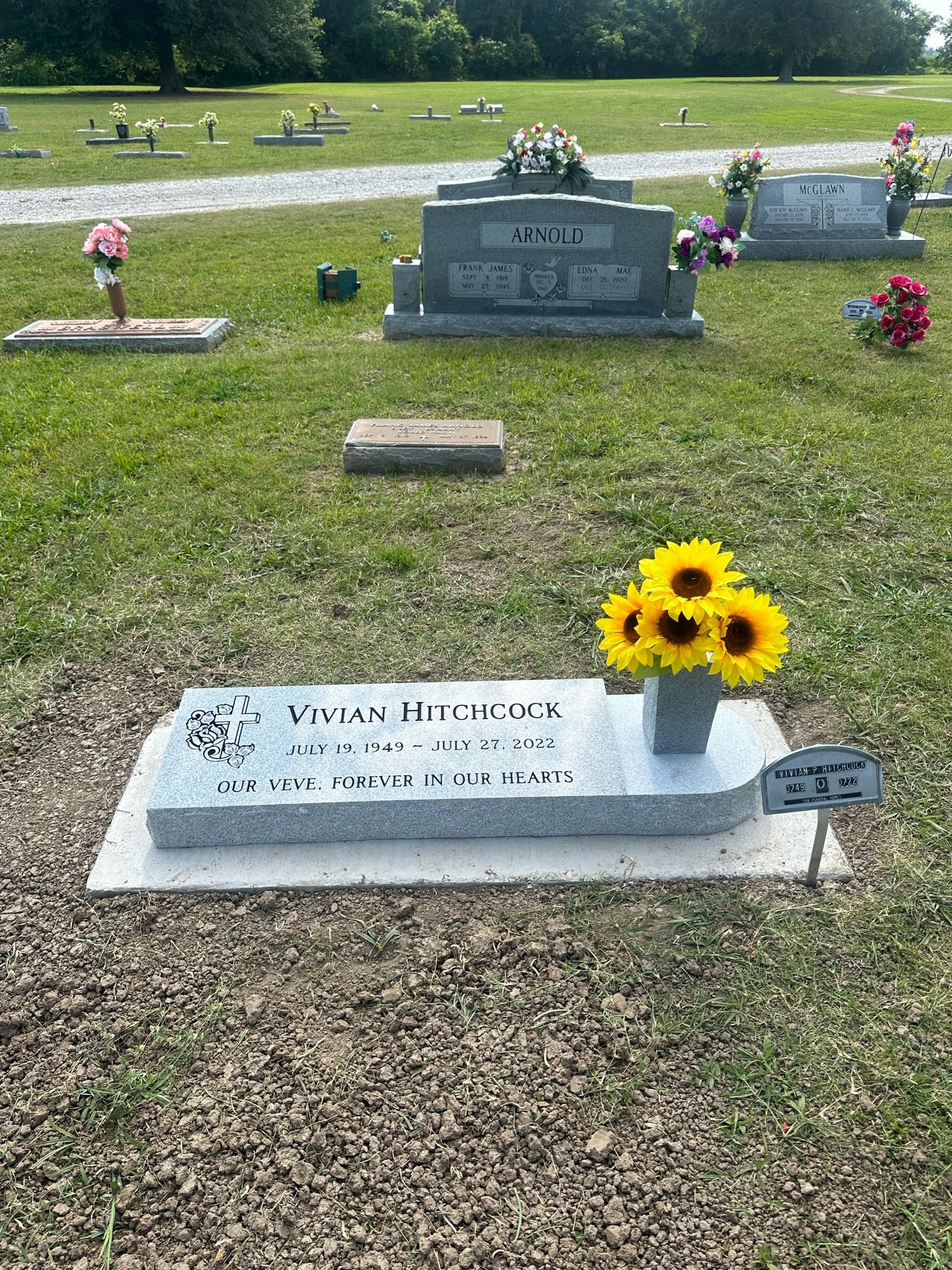 Grave of Vivian Hitchcock with a gray stone headstone, a vase with sunflowers, and a small plaque, in a cemetery with various other headstones and flowers in the background.