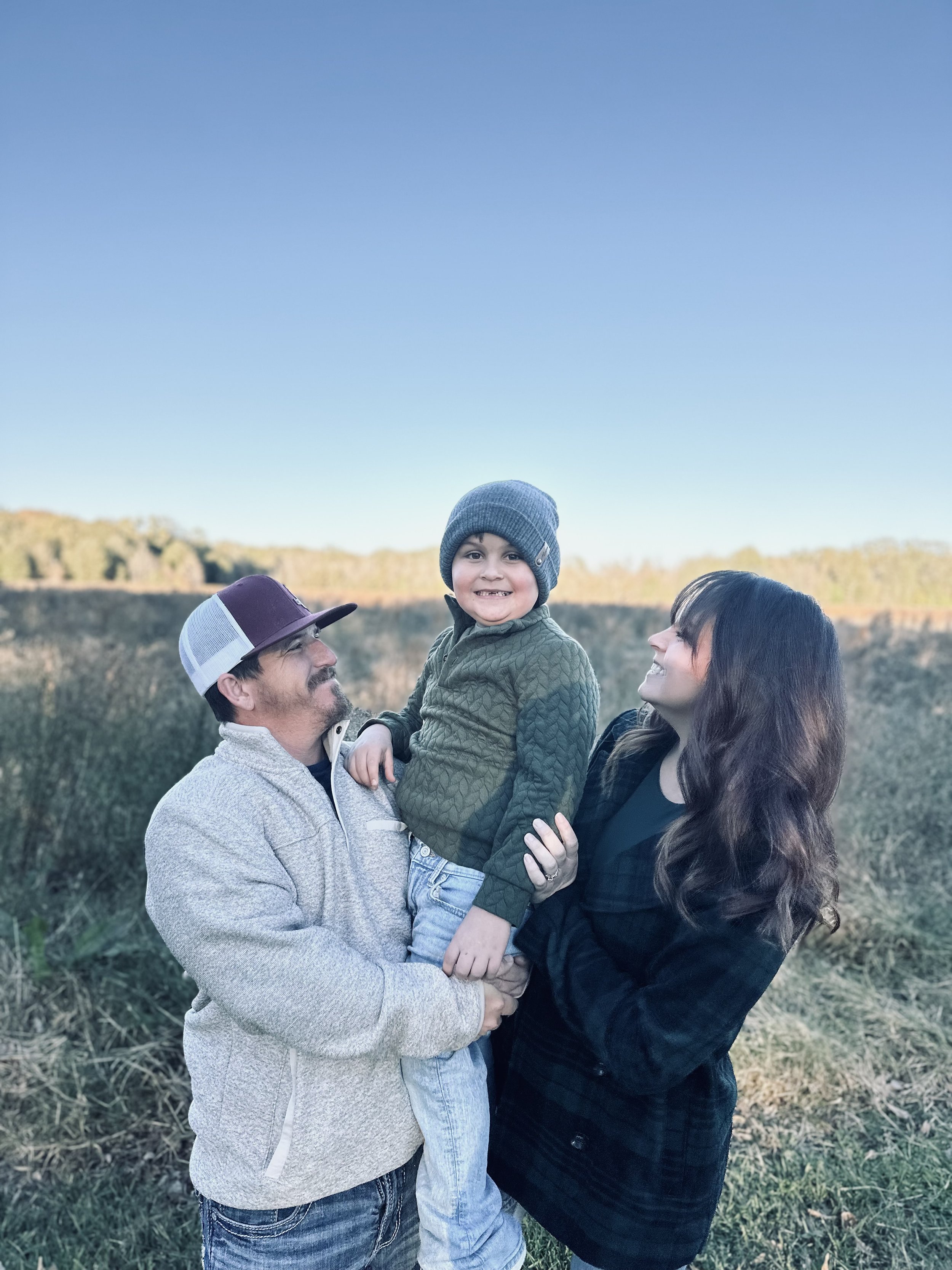 A smiling young boy in a gray beanie and a green quilted jacket is being held by a man wearing a white and purple cap and a light gray fleece jacket, while a woman with long dark hair and a black jacket looks on happily. They are outdoors in a field with clear blue sky in the background.