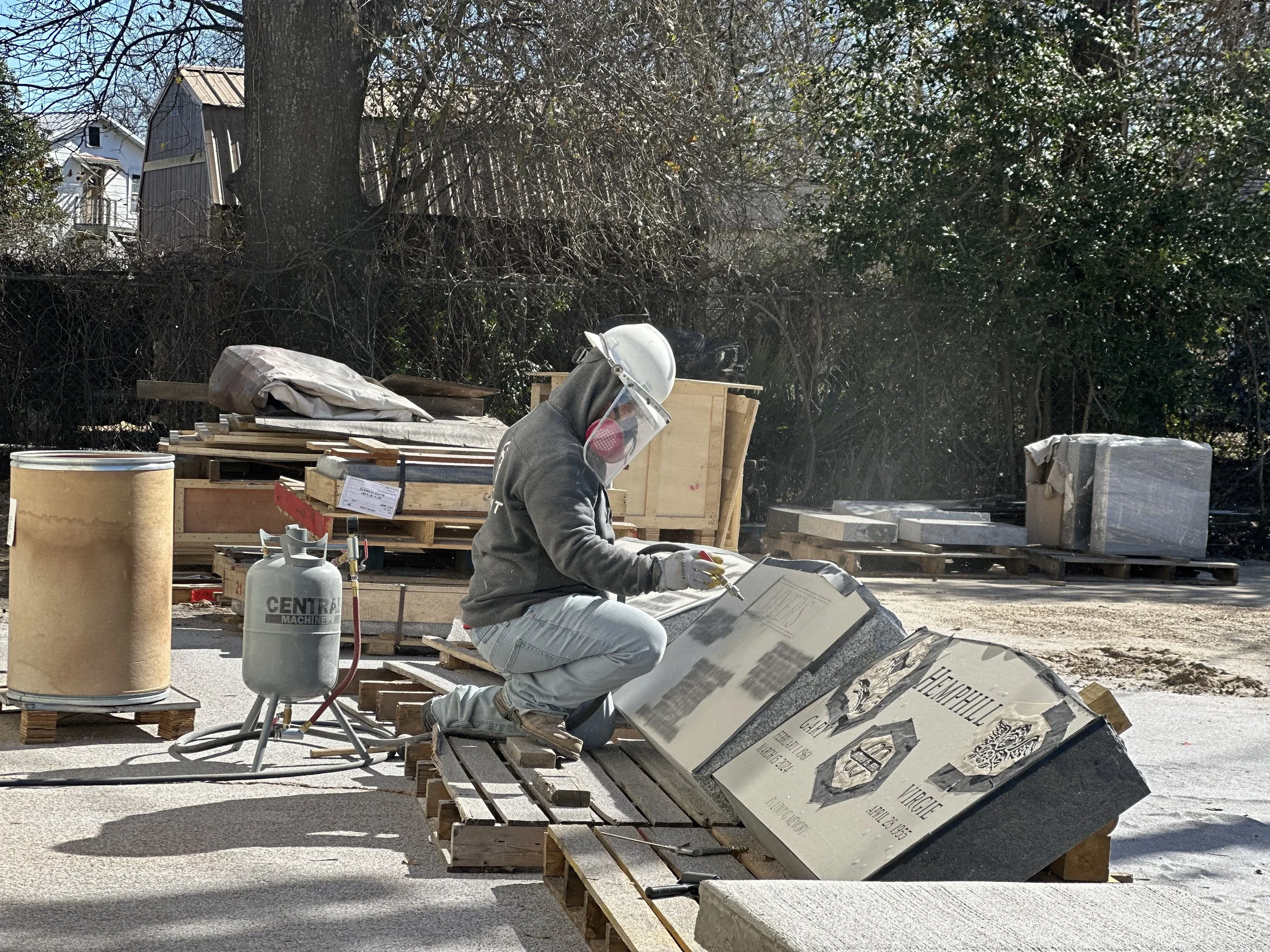 A worker kneeling on the ground in a construction or stone-working site, wearing a face shield and gloves, using a tool on a large stone plaque that reads 'Memorial' with additional text and images.