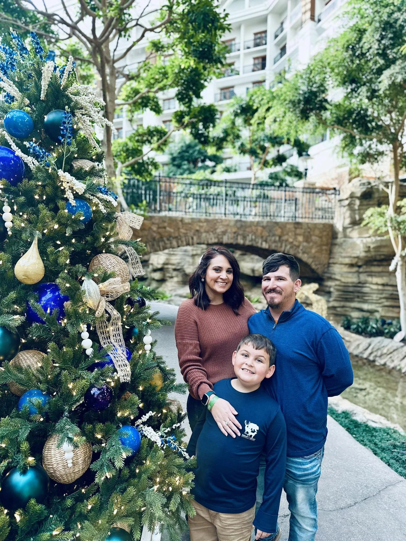 Family of three posing beside a decorated Christmas tree outdoors in a park area near a small pond, with trees, stone bridge, and apartment buildings in the background.