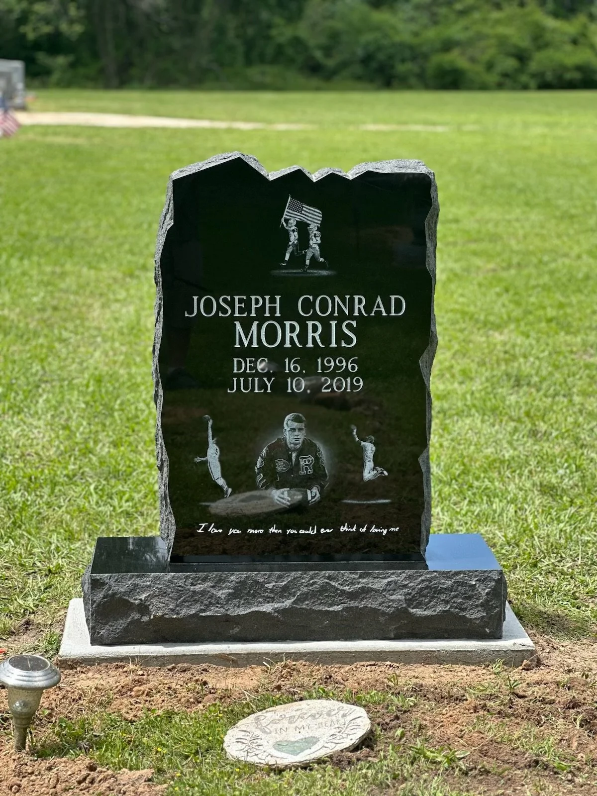 Black granite headstone with engraved images of a young man, some athletes, and holding an American flag. The headstone has the name Joseph Conrad Morris, born December 16, 1996, and died July 10, 2019. There is an inscription at the bottom that says