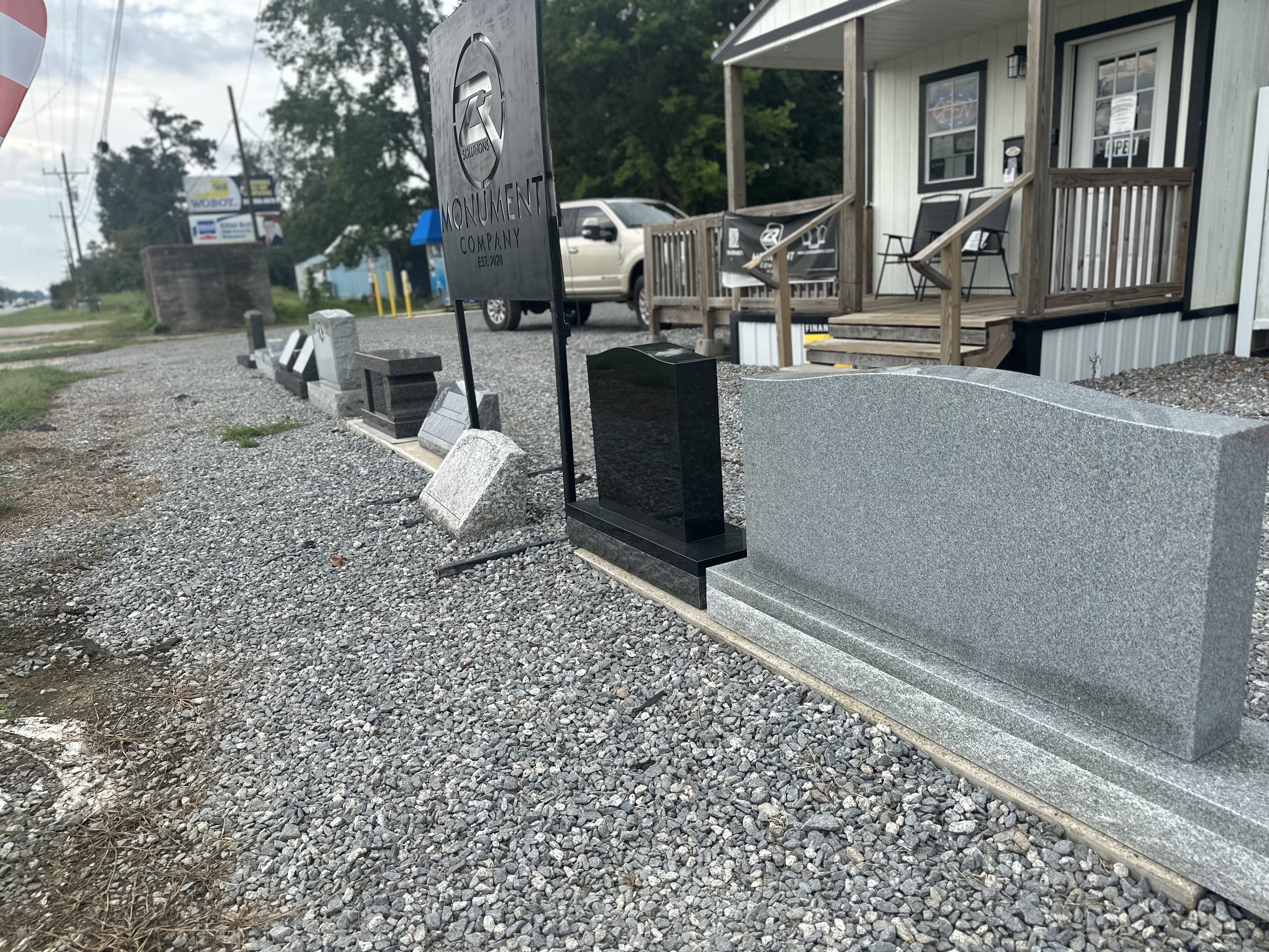 A row of different headstones with a sign that reads 'Monument Solutions' in front of a building with a porch and chairs.