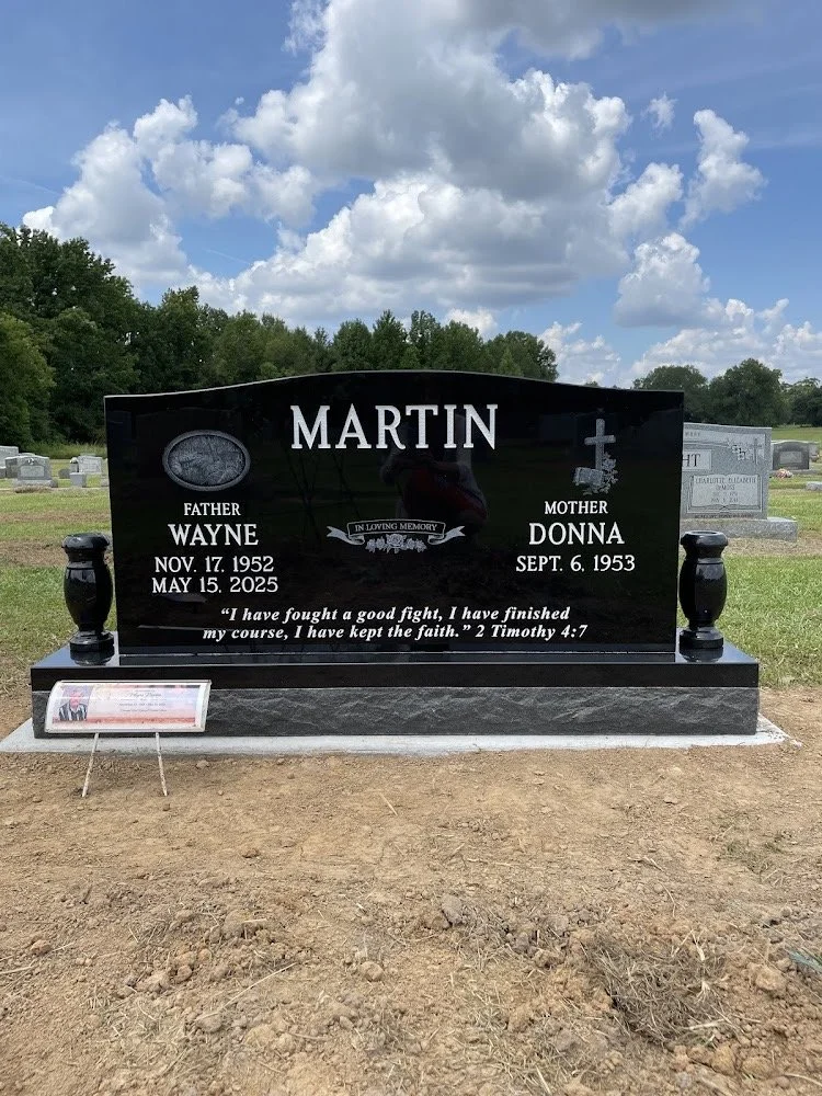 A gravestone for Martin with the names Wayne and Donna, birth and death dates, and a Bible verse from 2 Timothy 4:7. The gravestone is black with white lettering and is situated in a cemetery with other headstones in the background.