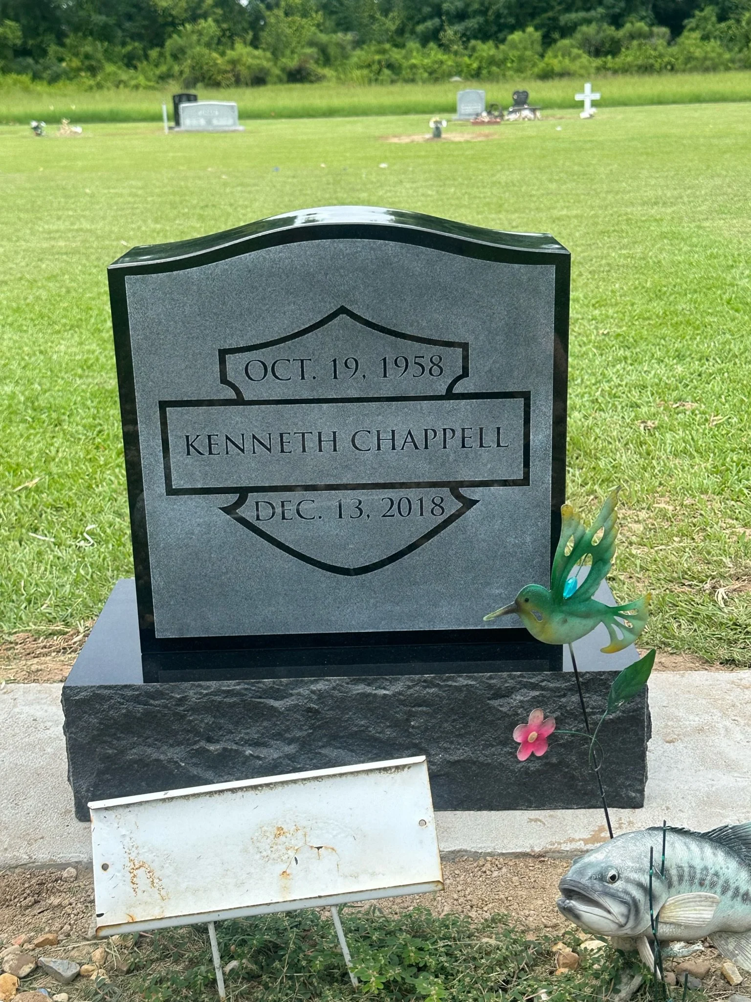 Gravestone with the name Kenneth Chappell, with dates October 19, 1958 and December 13, 2018, in a cemetery with other headstones and greenery in the background, decorated with colorful fishing-themed ornaments.