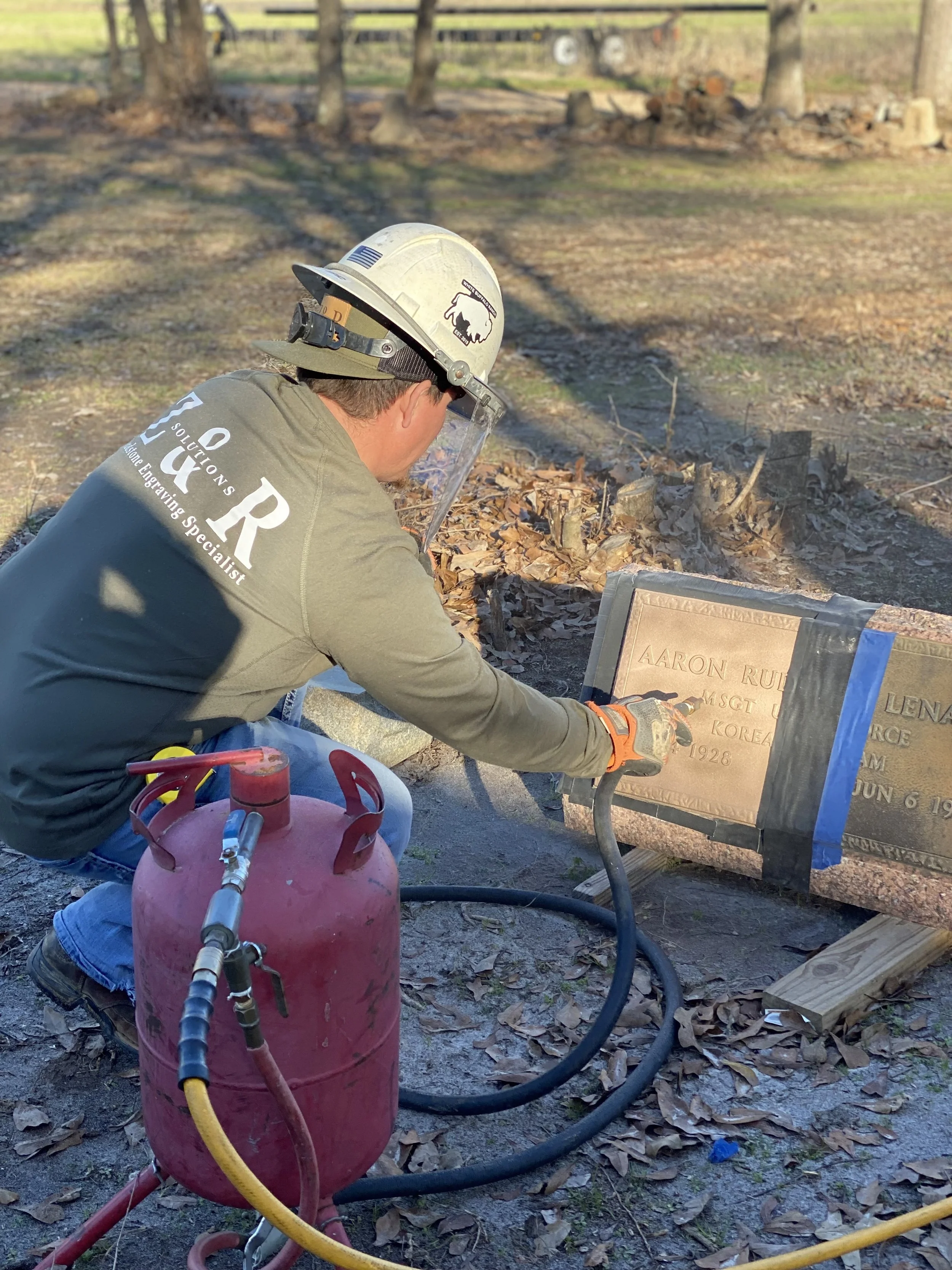 A worker wearing a helmet and face shield engraves a memorial plaque outdoors with a tool, with leaves on the ground and trees in the background.