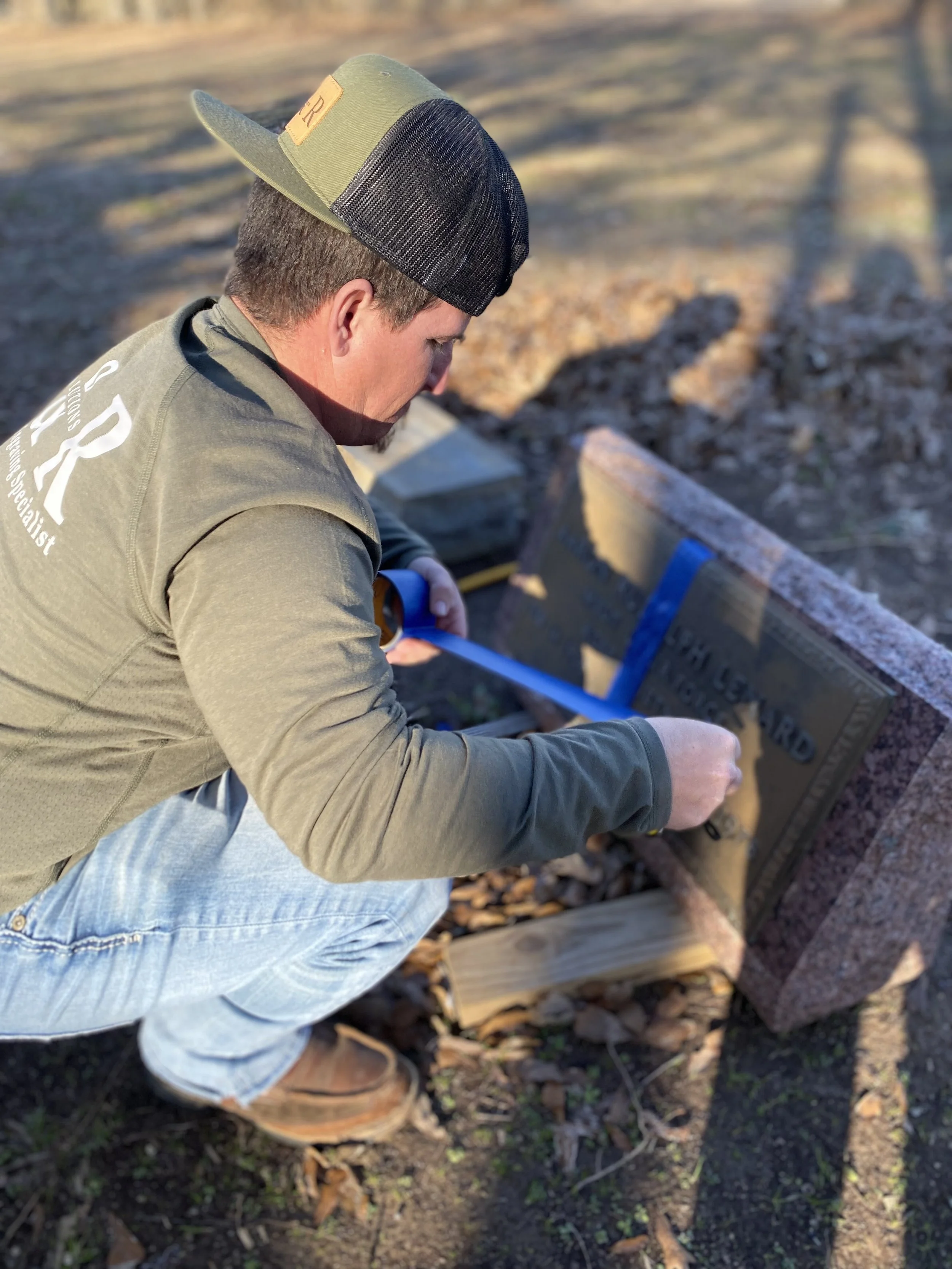 A man kneeling down outdoors, aligning a blue measuring tape across a granite marker with an inscription. He's wearing a green cap with a black mesh back and a tan jacket with the word 'ARK' on the back, and jeans. The background shows bare ground an