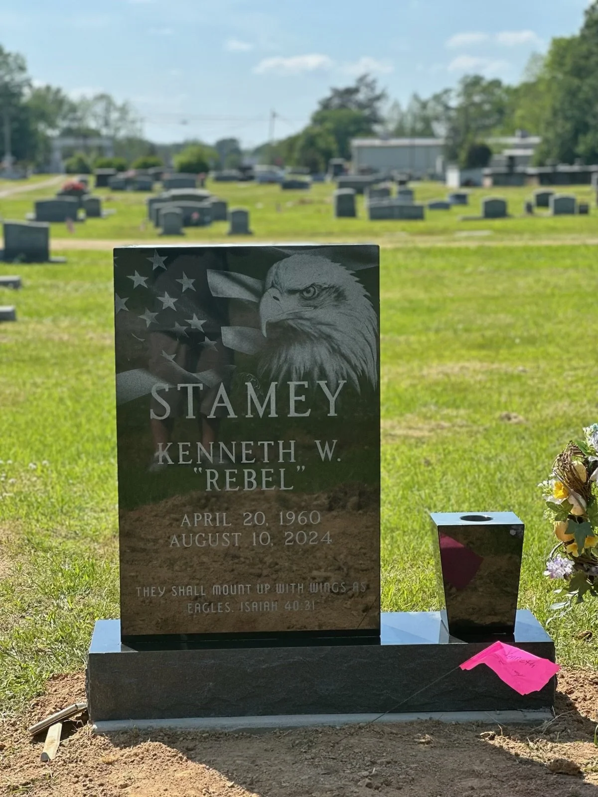 A black granite gravestone with an engraved eagle and American flag, dedicated to Kenneth W. 'Rebel' Stamey, with birth and death dates, located in a cemetery with many headstones and a cloudy sky.