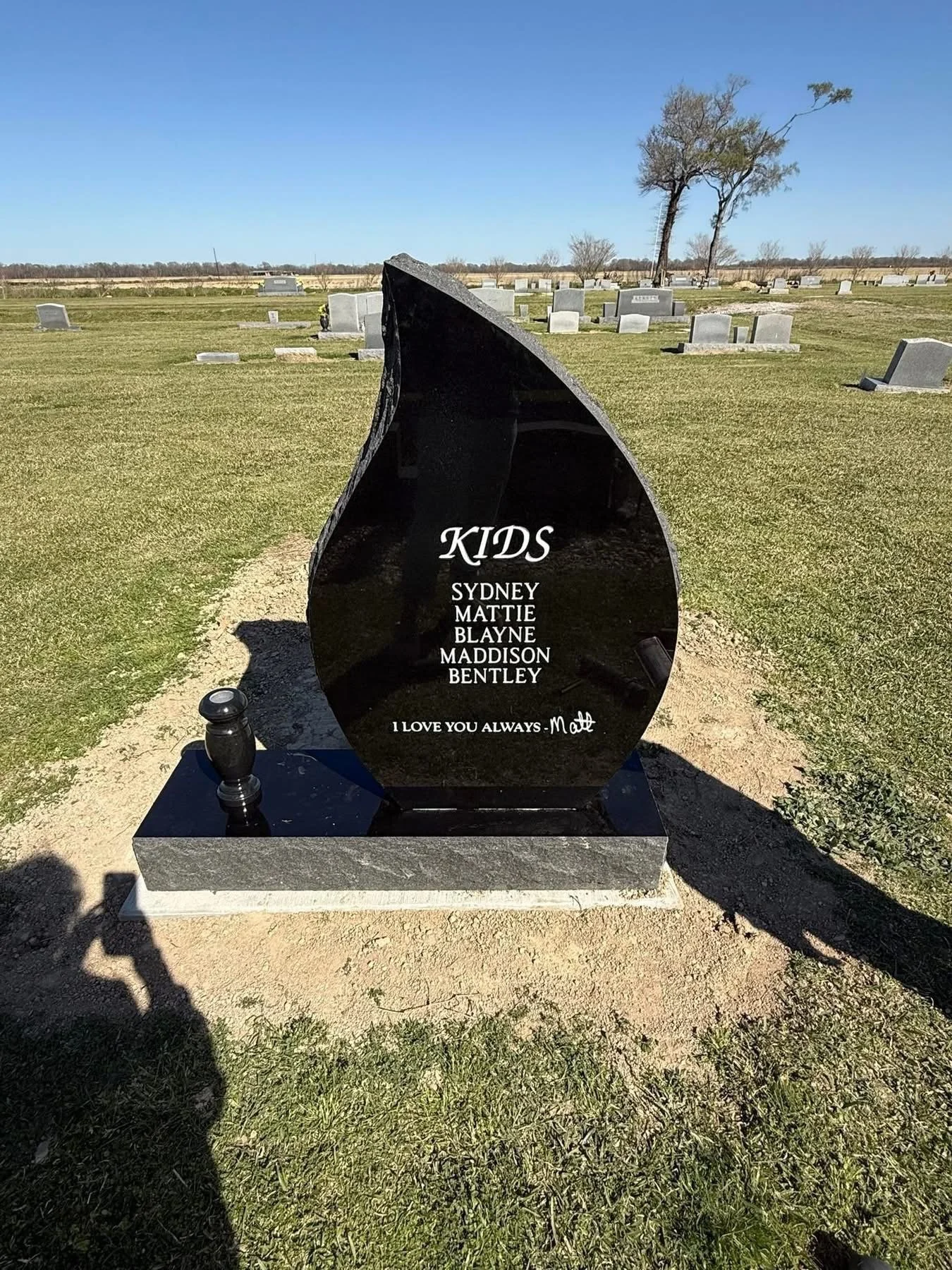 Black headstone at a cemetery with the word 'Kids' and a list of five names: Sydney, Mattie, Blayne, Maddison, Bentley. Below the names is a message: 'I love you always - Matt'.