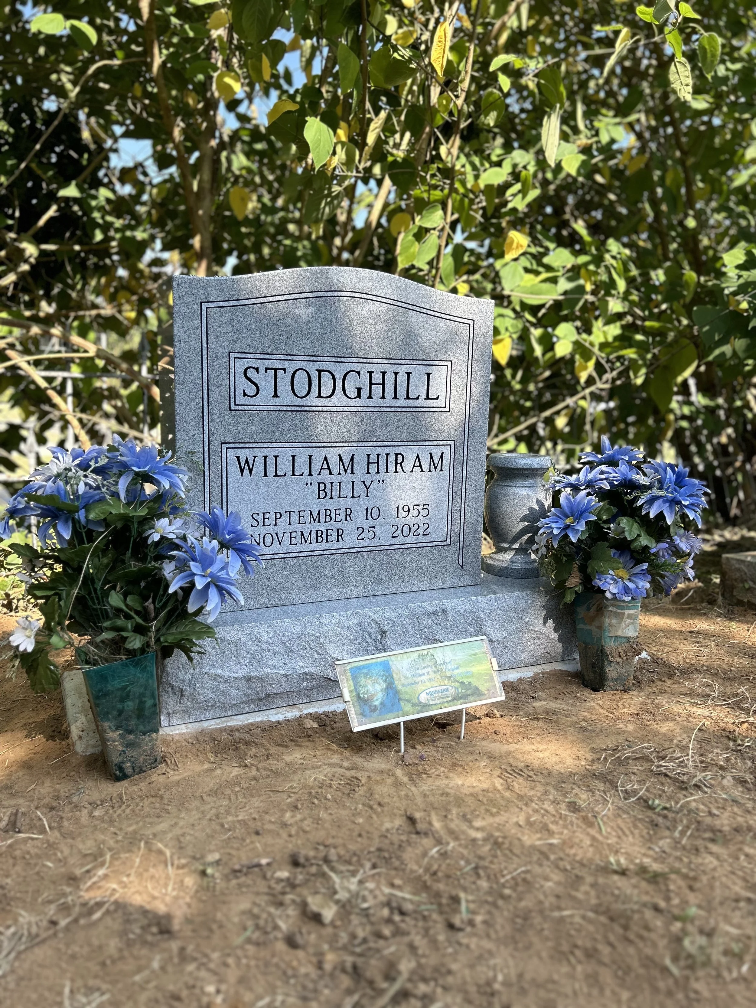Gravestone for William Hiram 'Billy' Stodghill, with blue flowers on either side, located outdoors with trees and foliage in the background.