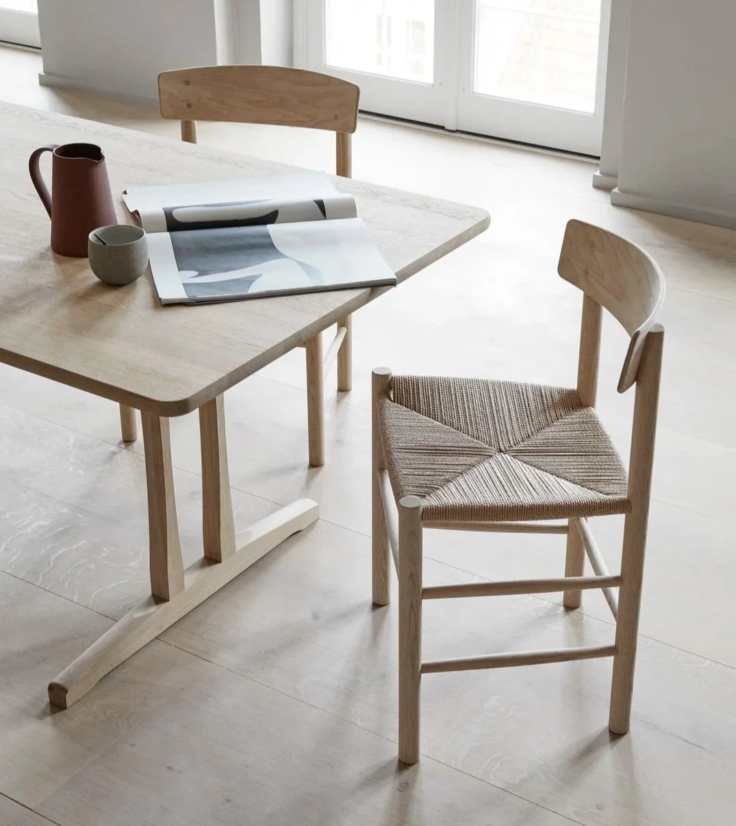 A wooden dining table with a magazine, a red pitcher, and a small grey bowl placed on it, accompanied by two light wood chairs in front of large windows with white frames, letting in natural daylight.