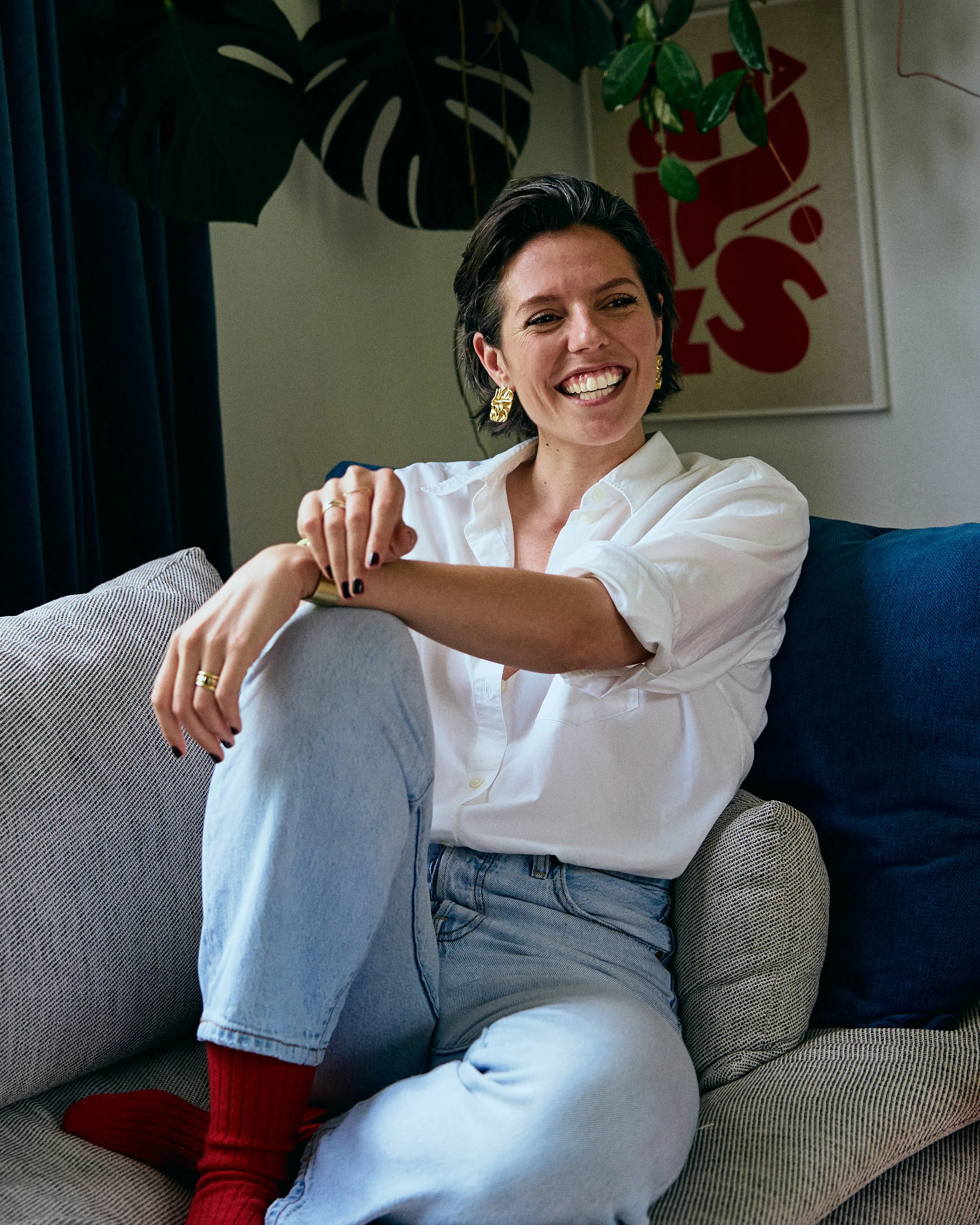 Woman with short brown hair, white blouse and gold earrings sitting on couch with arms crossed over knee and smiling friendly in front of abstract red art and monstera leaf