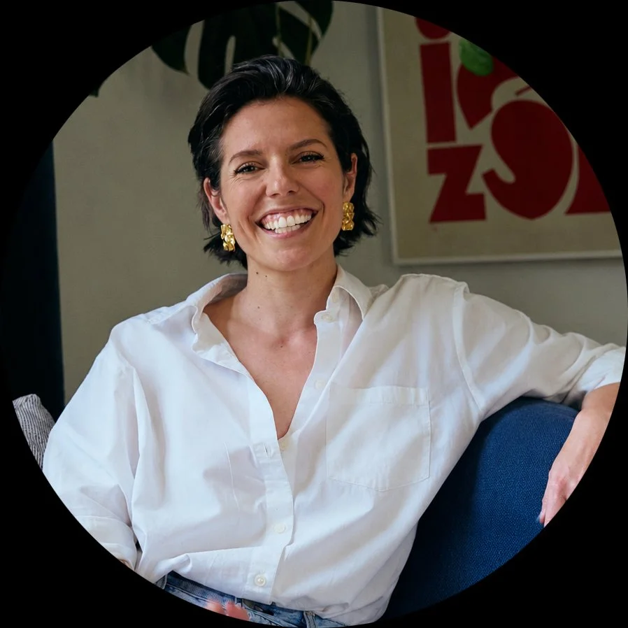 Woman with short brown hair, white blouse and gold earrings smiling invitingly sitting on blue couch in front of abstract art and monstera leaf