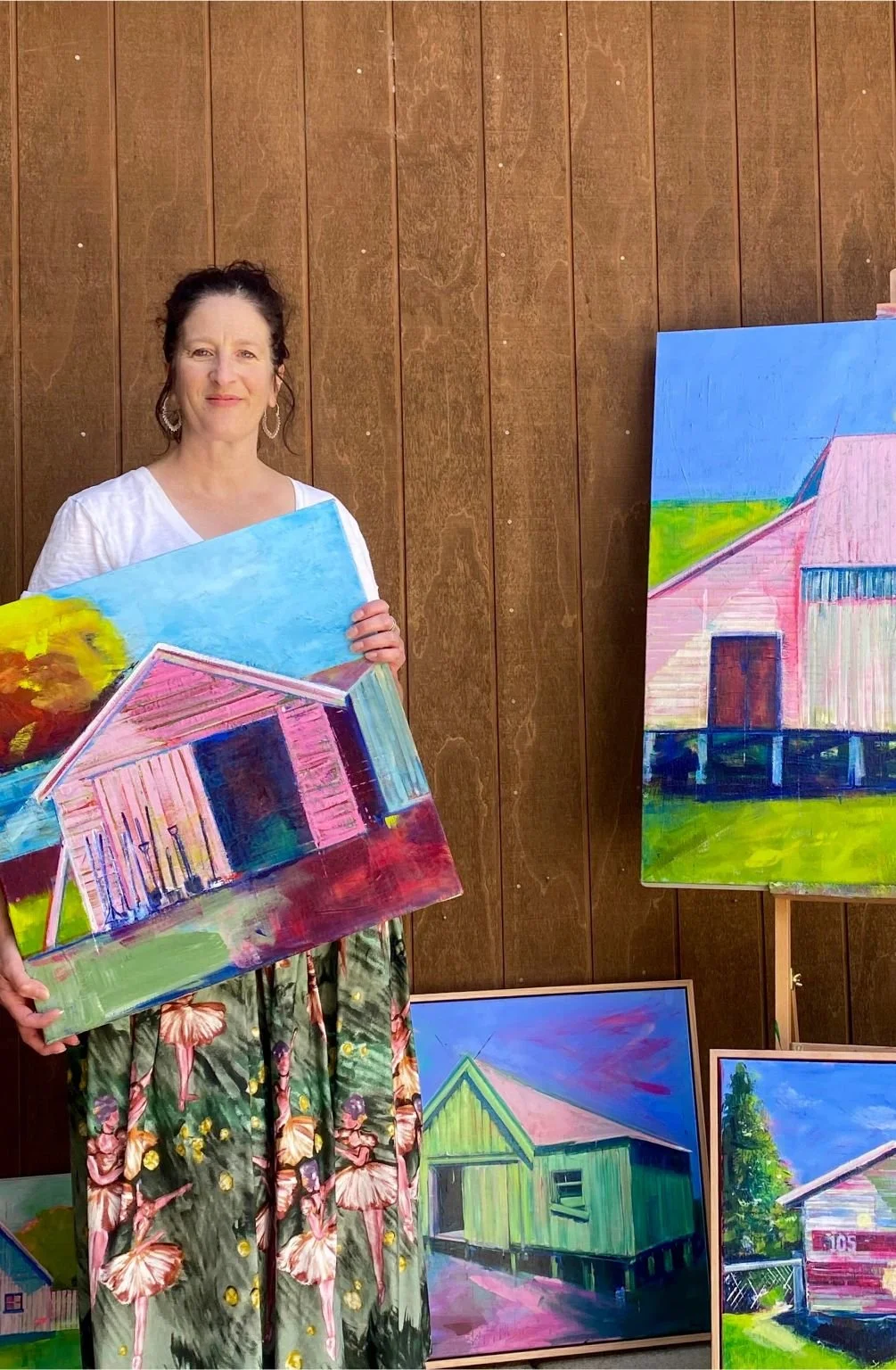 Artist Brooke Whelan stood in front of a wall holding a bright painting of a house.