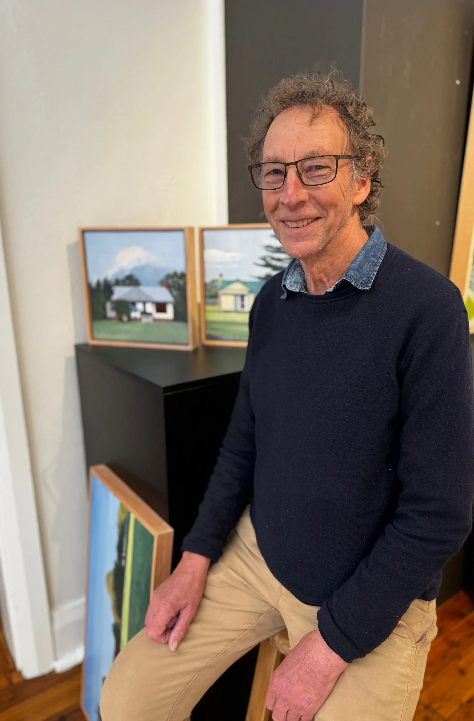 Artist Alan Harding seated on a stool in front of his paintings.