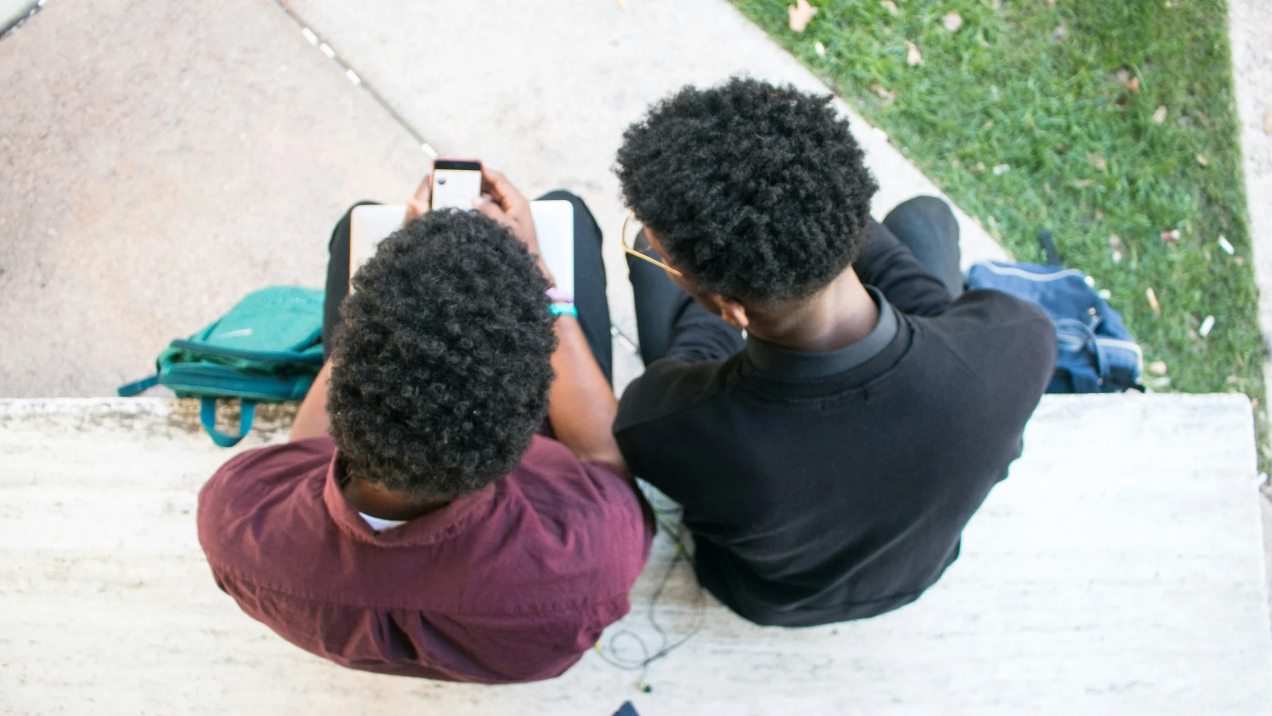 Overhead view of two Black teen boys, sitting outside, looking at a cell phone together.